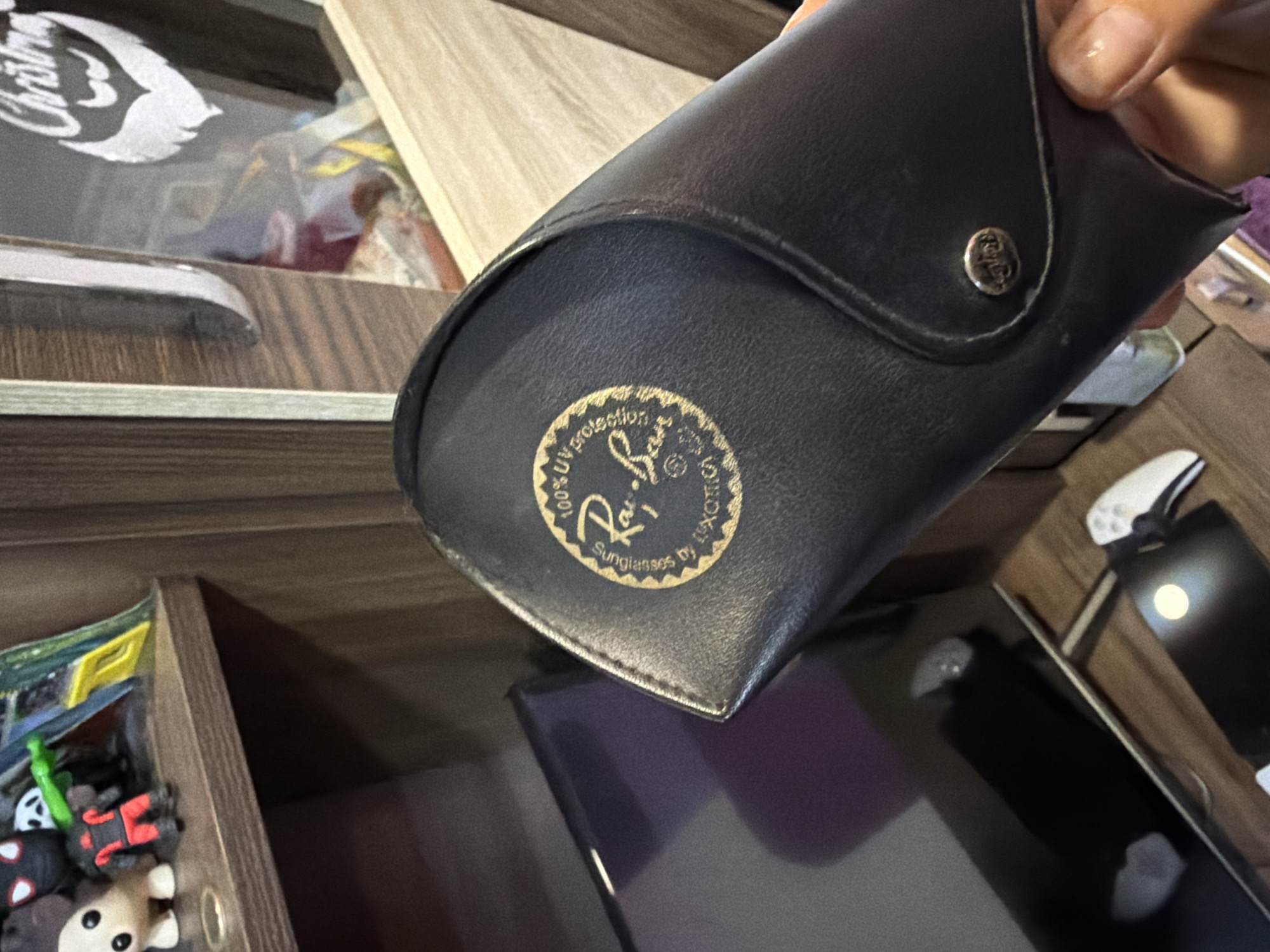 A close-up captures a dark leather Ray-Ban sunglasses case, featuring the brand's logo in gold print. The case is situated indoors, resting on a surface above an open shelf containing various small toys or figures. Part of a hand is visible holding the case, and in the background, a smartphone or tablet lies on a surface. The location is undefined.