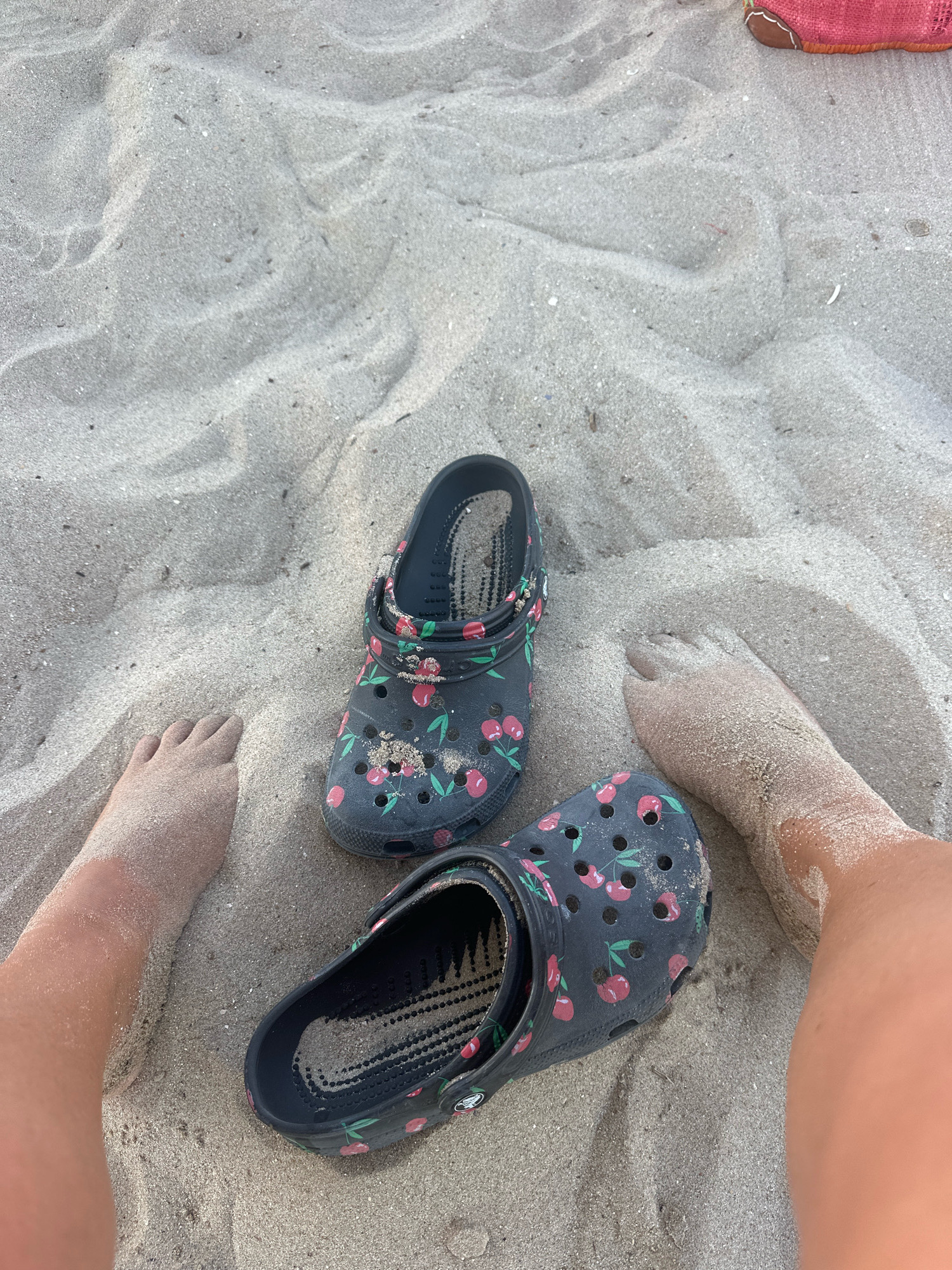 The image shows a pair of dark blue Crocs with a cherry pattern, resting on fine sand. One of the Crocs is partially filled with sand. Two feet, covered in sand, are visible in the foreground, suggesting the subjects are at a beach. The sandy ground is characterized by rippled textures. A hint of a pink bag is visible in the upper right corner. The scene is likely situated on the coast of Civitanova Marche, Italy, a city known for its beaches.