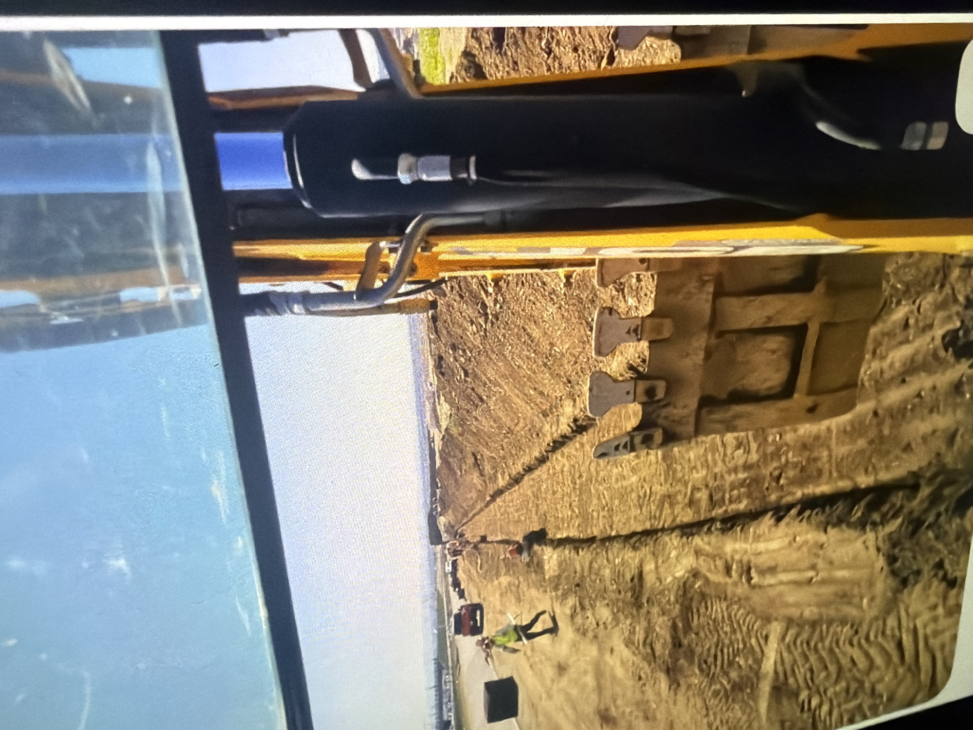 A wide-angle shot from the perspective of a construction vehicle shows a trench being dug in a dirt landscape. In the foreground, the yellow arm and bucket of the vehicle are visible, with metal teeth on the bucket. In the midground, a worker in a yellow vest and hard hat is walking away from the camera, towards a red truck parked on a dirt road. A trench runs parallel to the road, with piles of dirt on either side. The background shows a hazy horizon with distant buildings and a clear blue sky. The overall scene depicts an active construction site.