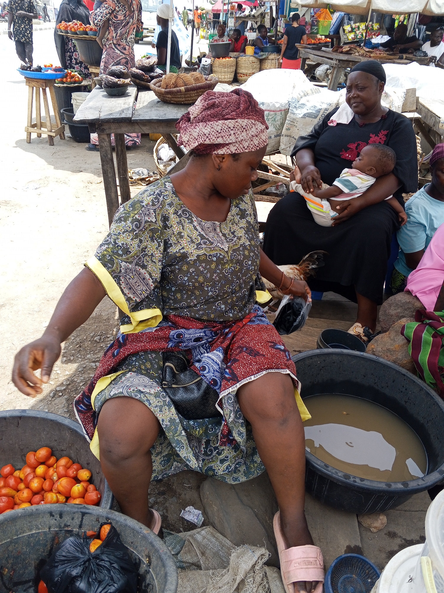The image captures a scene in Zango-ndaji, Nigeria, showing a marketplace environment. A woman wearing a patterned dress and headwrap sits with a bucket of tomatoes beside her. Another woman in a black dress is seated holding a baby. A chicken is visible near the woman in the black dress. A bucket of water sits next to the woman in the patterned dress. Other vendors and customers are visible in the background, along with tables displaying various goods.
