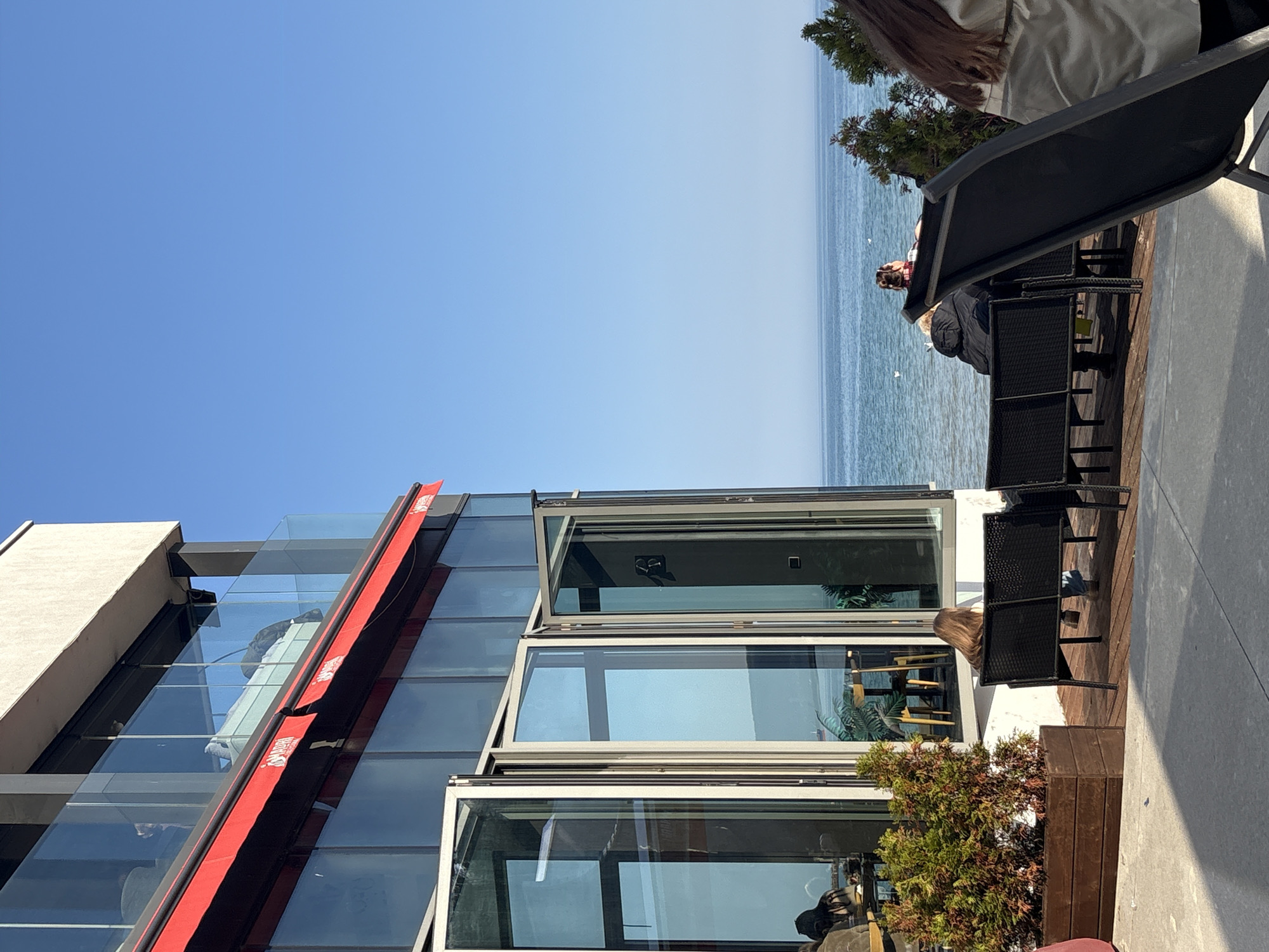 The image shows an outdoor patio area with a modern building next to a body of water, likely the Black Sea. The building has large glass windows, some of which are open. A red awning runs along part of the building. People are seated at black patio chairs, some facing the water. There are also some plants in planters. The sky is blue. Given the location on the Black Sea, it's possible this scene is in Varna, Bulgaria. More information is needed to confirm.