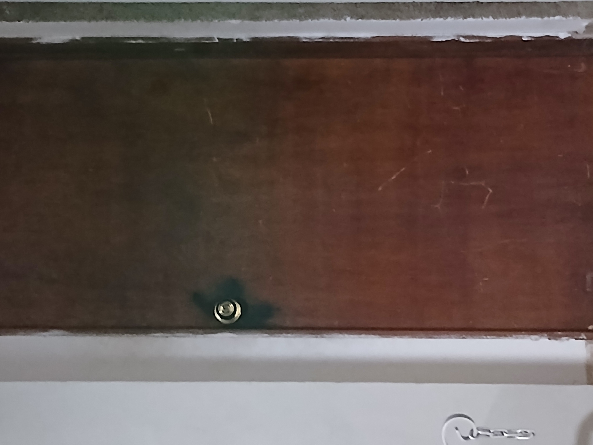A wooden door, a wall, and a key hook are visible in this image. The door is a dark brown color with some scuff marks and scratches on its surface. It has a round, metallic doorknob, which appears to be a silver-tone. To the left of the door, a white wall is present, with a metallic key hook attached to it. The key hook consists of a circular loop from which a chain hangs down. The scene appears to be indoors, likely in a residential or commercial setting. The image does not provide information about the specific location within Santa Ana, Venezuela.