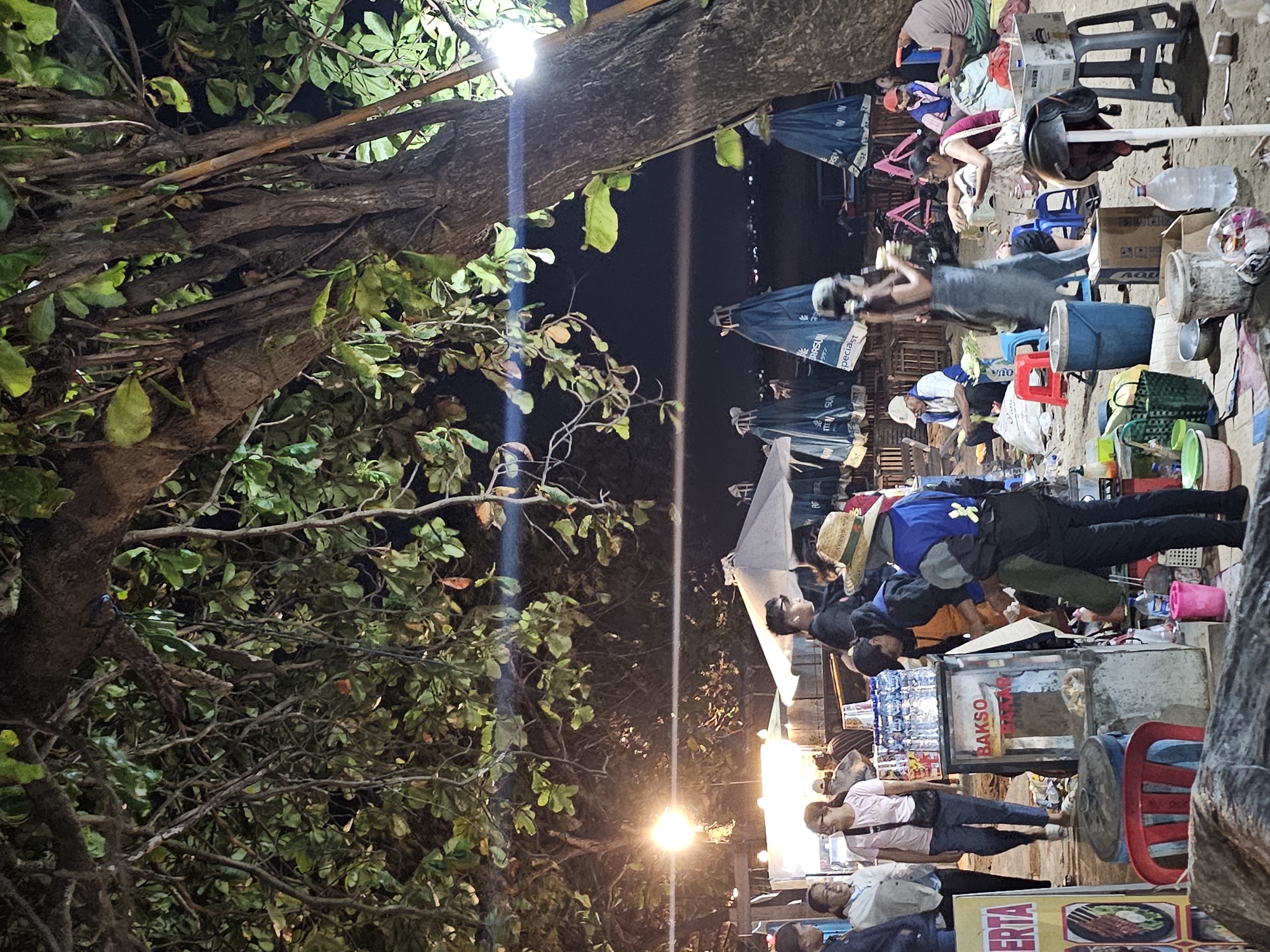 This image captures a bustling night scene on a beach, likely in Denpasar, Indonesia, given the presence of food stalls and local people. In the foreground, a large tree with dense foliage dominates the right side of the frame, its branches casting shadows across the scene.
Several food stalls are set up, illuminated by bright lights. One stall prominently displays "BAKSO" in red lettering, indicating meatballs are being sold. Bottles of water are stacked neatly on top of this stall. Various individuals are present, some appearing to be vendors and others patrons.
In the mid-ground, people are gathered around the stalls, some eating, some talking, and some appearing to be ordering food. There are umbrellas set up, some closed and some open, suggesting a beachside marketplace.
The ground is sandy, consistent with a beach environment, and scattered with various items including buckets, baskets, plastic containers, and chairs. In the background, more umbrellas and what appear to be beach chairs are visible, and a body of water, likely the ocean, can be discerned under the night sky. The overall atmosphere is lively and suggests a typical evening gathering at a popular beach location.