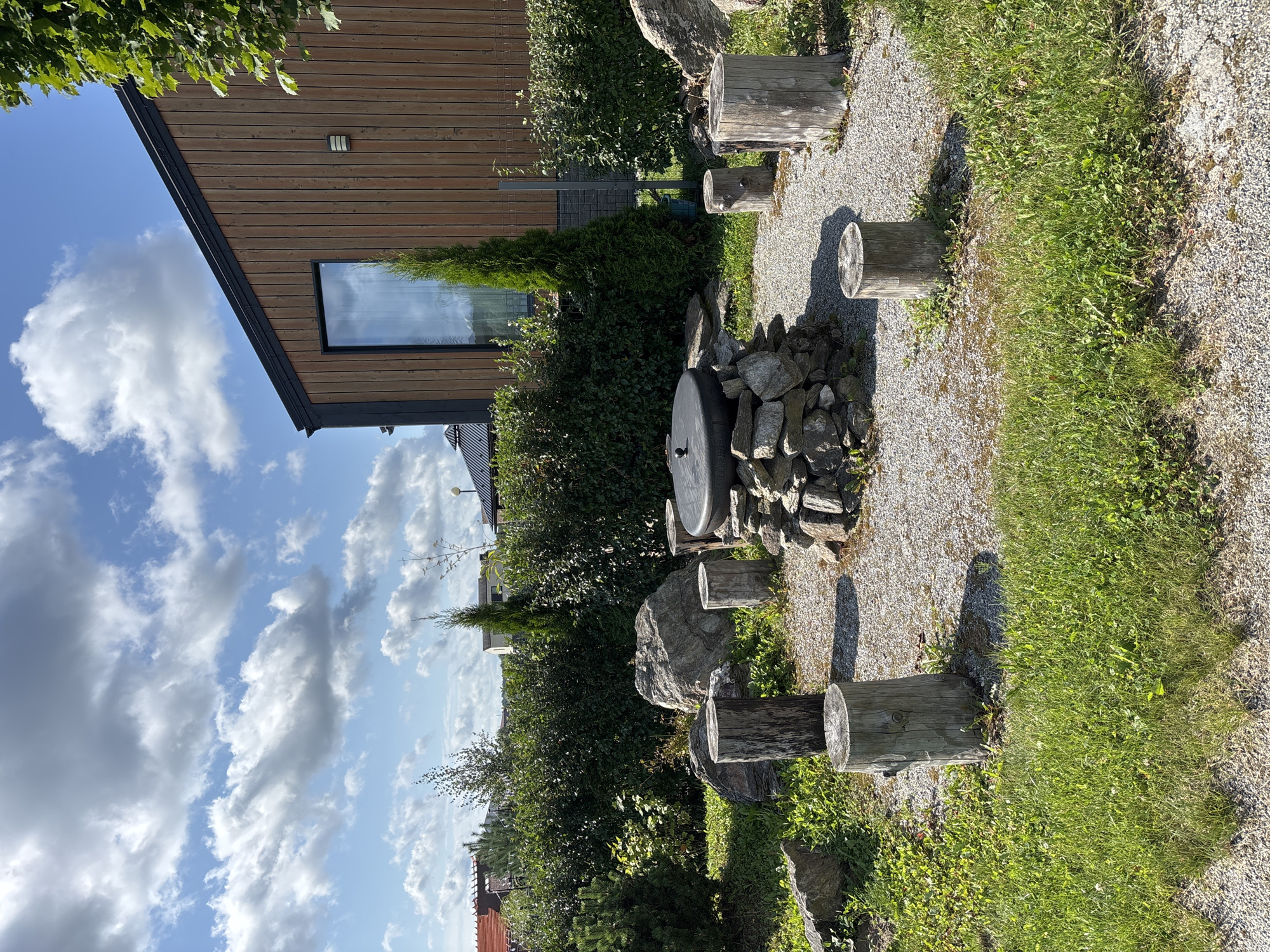 A natural outdoor seating area is depicted, featuring a central stone fire pit with a dark, circular lid. Surrounding the fire pit are several tree stump seats, some arranged in a rough circle. Large rocks are scattered around the area, adding to the natural aesthetic. A dense green hedge forms a backdrop, with a modern wooden building with a large window visible behind it. The ground is covered in gravel and patches of green grass. The sky above is blue with scattered white clouds. The scene appears to be in a residential area, possibly a backyard, in Zvenigorod, Russia. There are no people present.
