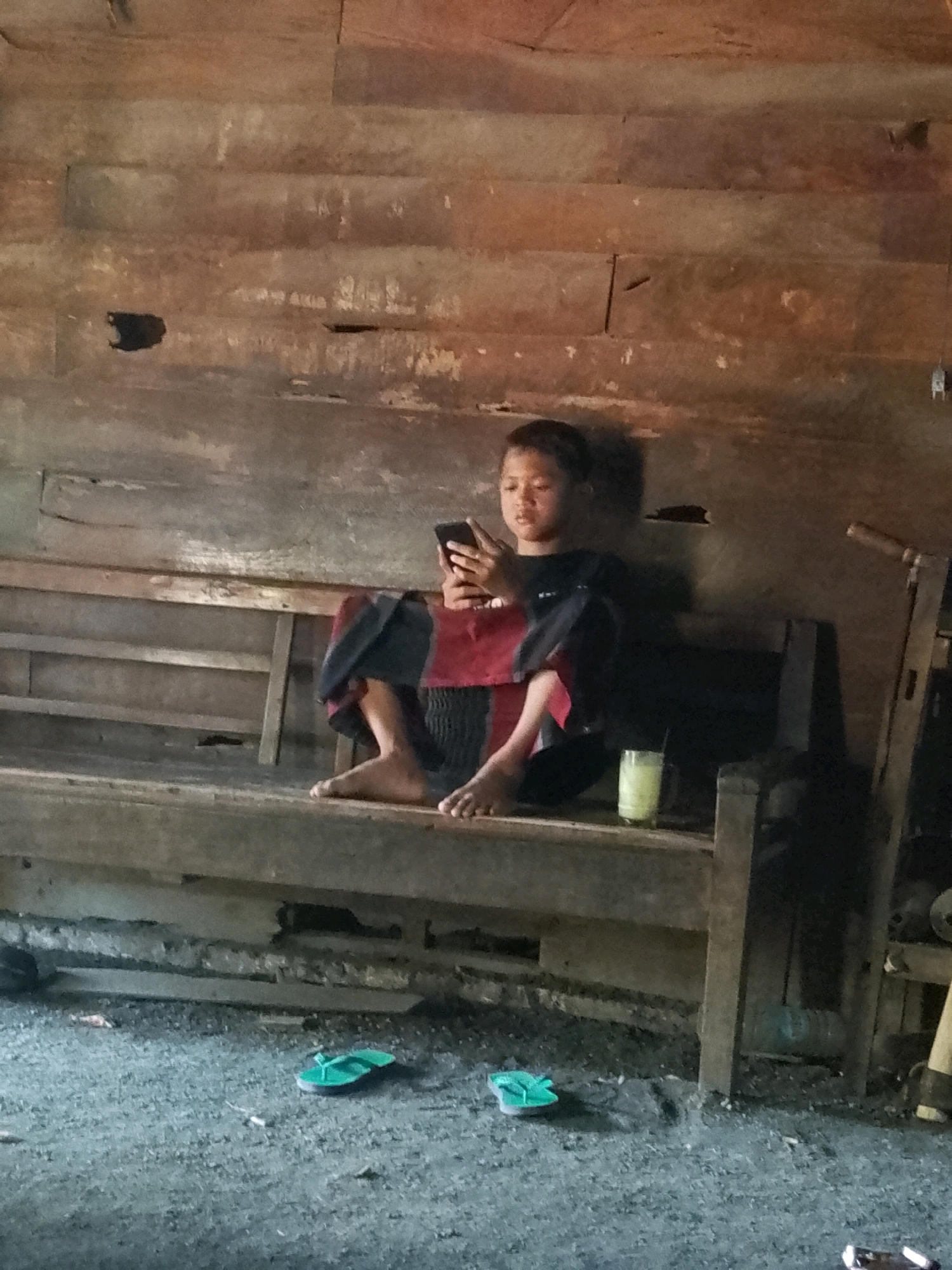A young person sits on a wooden bench in a rustic, unfinished wooden structure. The person is holding a smartphone in their right hand and appears to be looking at it. They are wearing a dark long-sleeved shirt and a red and black patterned cloth around their waist. A glass of pale yellow liquid with a straw is placed on the bench to the person's right. The floor is a rough, gritty surface. Two green flip-flops are visible on the floor in the foreground, one to the left and one to the right of the bench. The walls of the structure are made of horizontal wooden planks, some of which are weathered and appear damaged. The overall impression is one of a simple, possibly rural, dwelling. The location is Wadang, Indonesia.