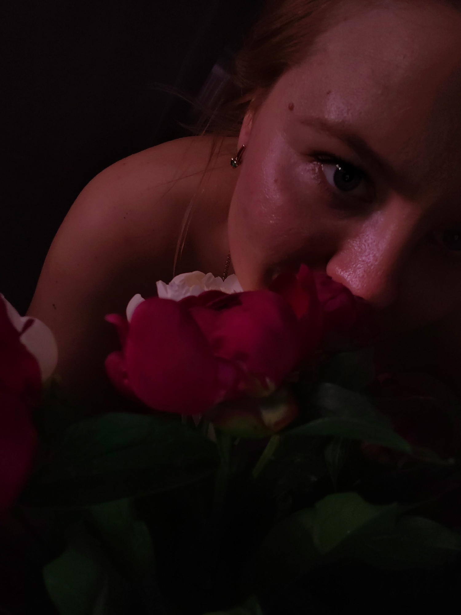 The image shows a close-up of a woman's face and shoulder, with a bouquet of peonies in the foreground. The woman's hair is light brown, and she is wearing a gold earring. Her skin is illuminated by a pinkish light, highlighting her features. The peonies are a deep pink and white, and their petals are soft and delicate. The background is dark and out of focus, creating a sense of intimacy and focus on the woman and flowers. There are no discernible landmarks or clues to indicate a specific location in Izhevsk, Russia, or any other city. The image is a still photograph, capturing a moment of quiet contemplation or appreciation of nature.
