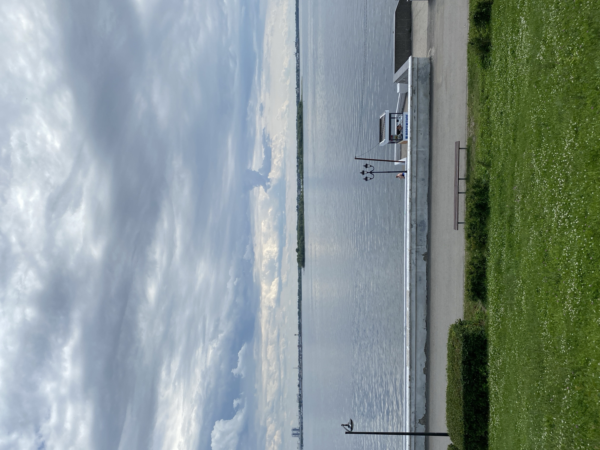 The image shows a wide, calm body of water under a cloudy sky. In the foreground, a lush green lawn dotted with small white flowers slopes down to a paved walkway. A neatly trimmed hedge runs along the edge of the walkway, beyond which is a concrete barrier topped with a white railing. A lamppost stands to the left. Further along the walkway, there is a small kiosk with a sign that reads "МОРОЖЕНОЕ" (ice cream in Russian). A person is visible near the kiosk, and another person is seated on a wooden bench further down the walkway. Across the water, a long strip of land with trees and buildings is visible, with more distant structures on the horizon. The overall impression is a tranquil, somewhat overcast day by the water.