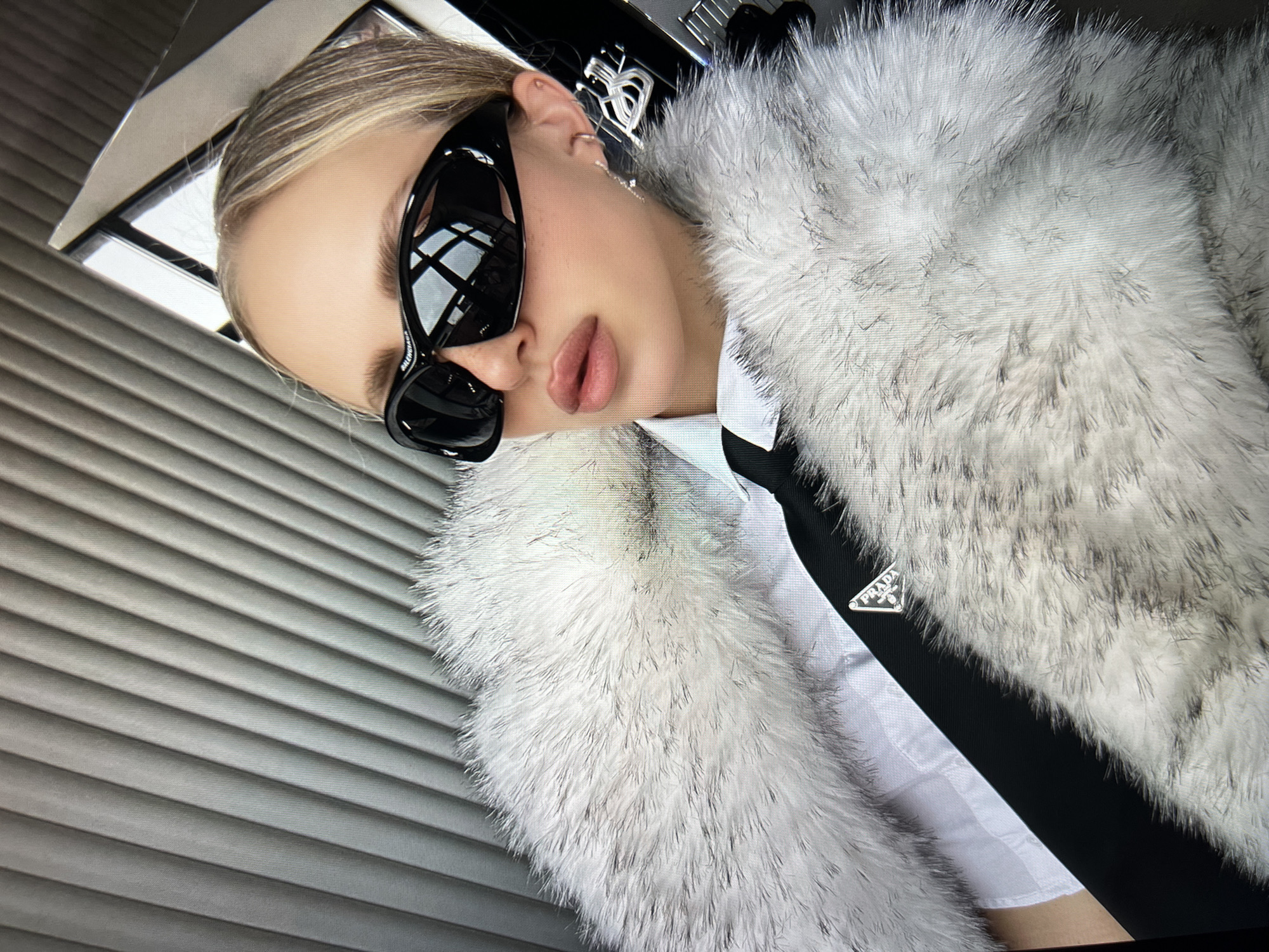 The image shows a close-up, high-angle shot of a woman wearing a white collared shirt, a black tie with a Prada logo, and a thick, fluffy white fur coat. She has blonde hair pulled back and is wearing large, black, wraparound sunglasses. Her lips are painted a pale pink. The background appears to be the interior of a vehicle, with a textured grey surface that could be a seat or wall paneling, and a window reflecting the sky and what appears to be an urban setting with modern architecture. The lighting suggests it is daytime. There is no indication of the city of Saint Petersburg, Russia, or any specific urban location.