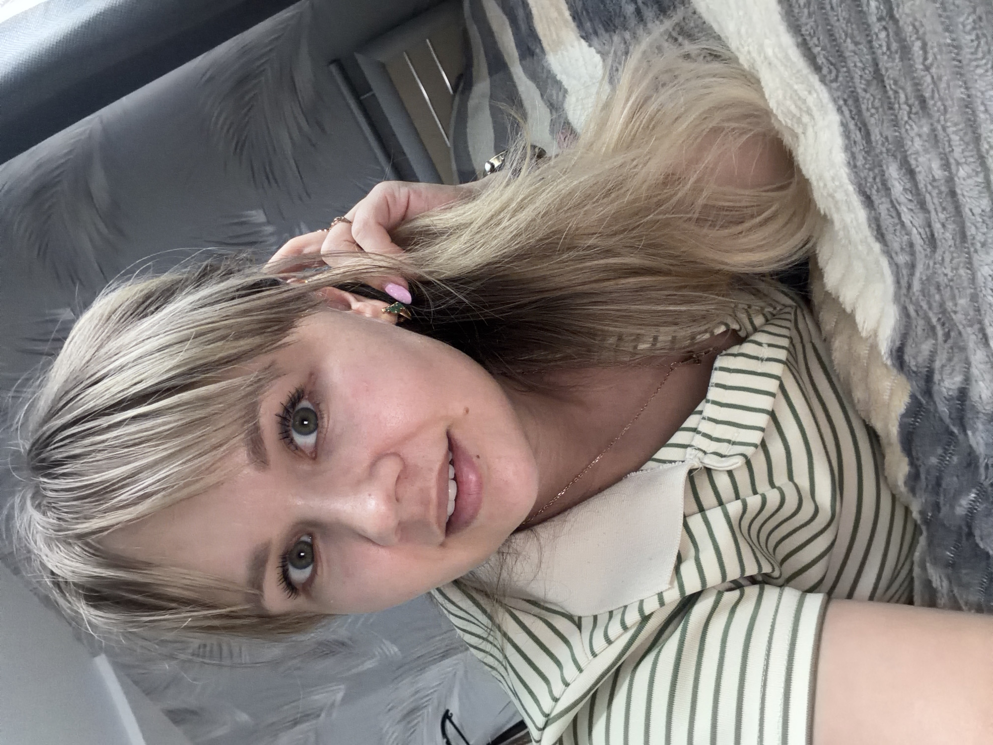 A close-up portrait shows a woman lying down, looking directly at the camera with a slight smile. She has long, blonde hair with bangs, and her eyes are light green. She is wearing a light-colored, possibly cream or off-white, collared shirt with thin green vertical stripes. A delicate gold chain necklace is visible around her neck. On her left hand, which is raised near her head, she has multiple rings on her fingers and her fingernails are painted pink. A small earring, possibly gold with a green stone, is visible in her left ear.
To her right, a striped blanket or throw in shades of grey and white with a fuzzy texture is partially covering her. The background behind her head appears to be a grey surface with a subtle, dark patterned design, possibly wallpaper or a headboard. The overall setting suggests a relaxed indoor environment, possibly on a bed or couch. The lighting is soft and even, indicating an indoor daytime scene. No text is visible in the image.