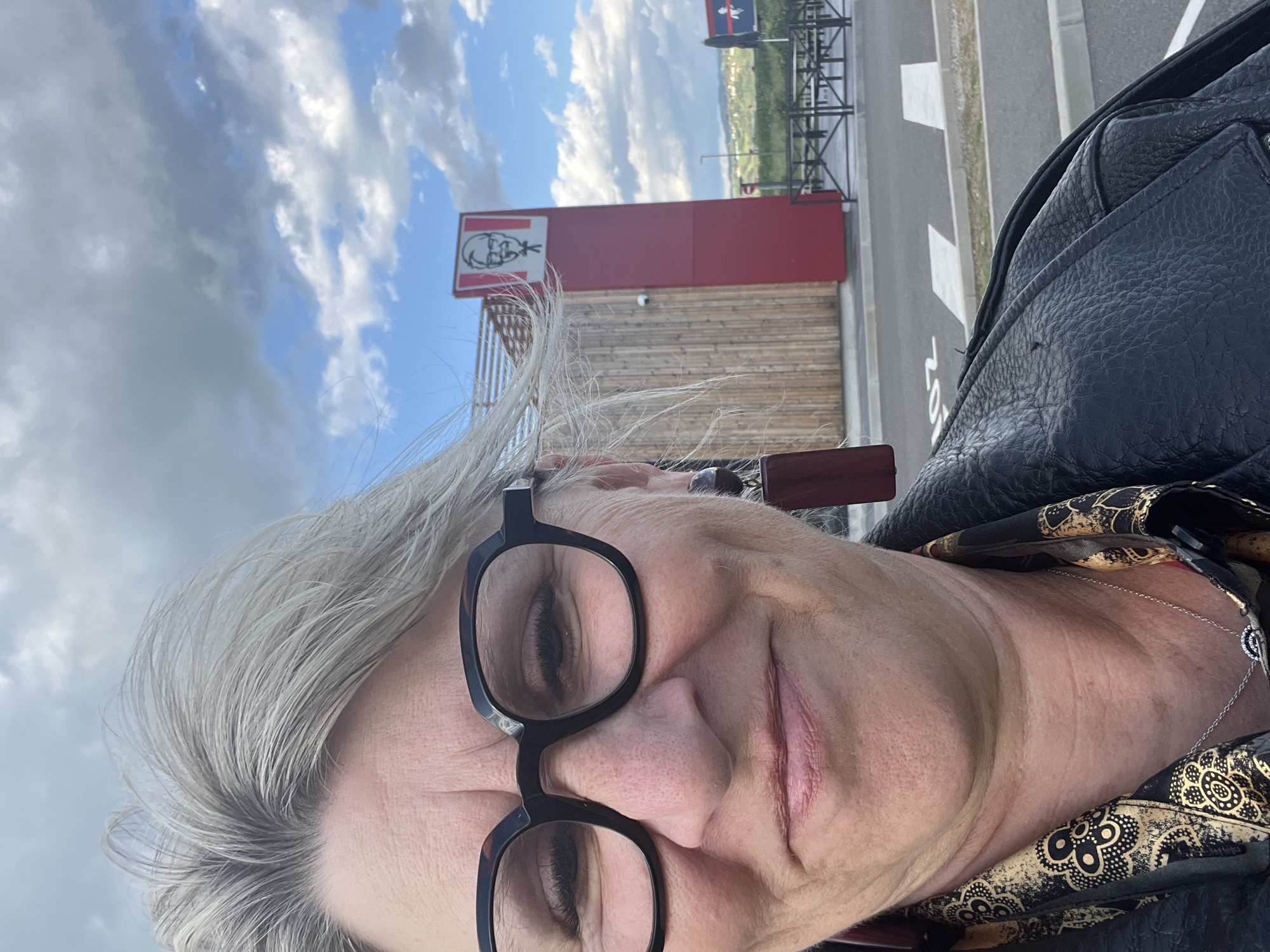 The image shows a selfie of a mature woman with short grey hair and glasses, wearing a black leather jacket and a patterned scarf with a silver necklace. In the background, a KFC restaurant is visible with its iconic logo and signage. The location appears to be in Andrézieux-Bouthéon, France, as per the metadata. A road and some greenery are also visible in the background.