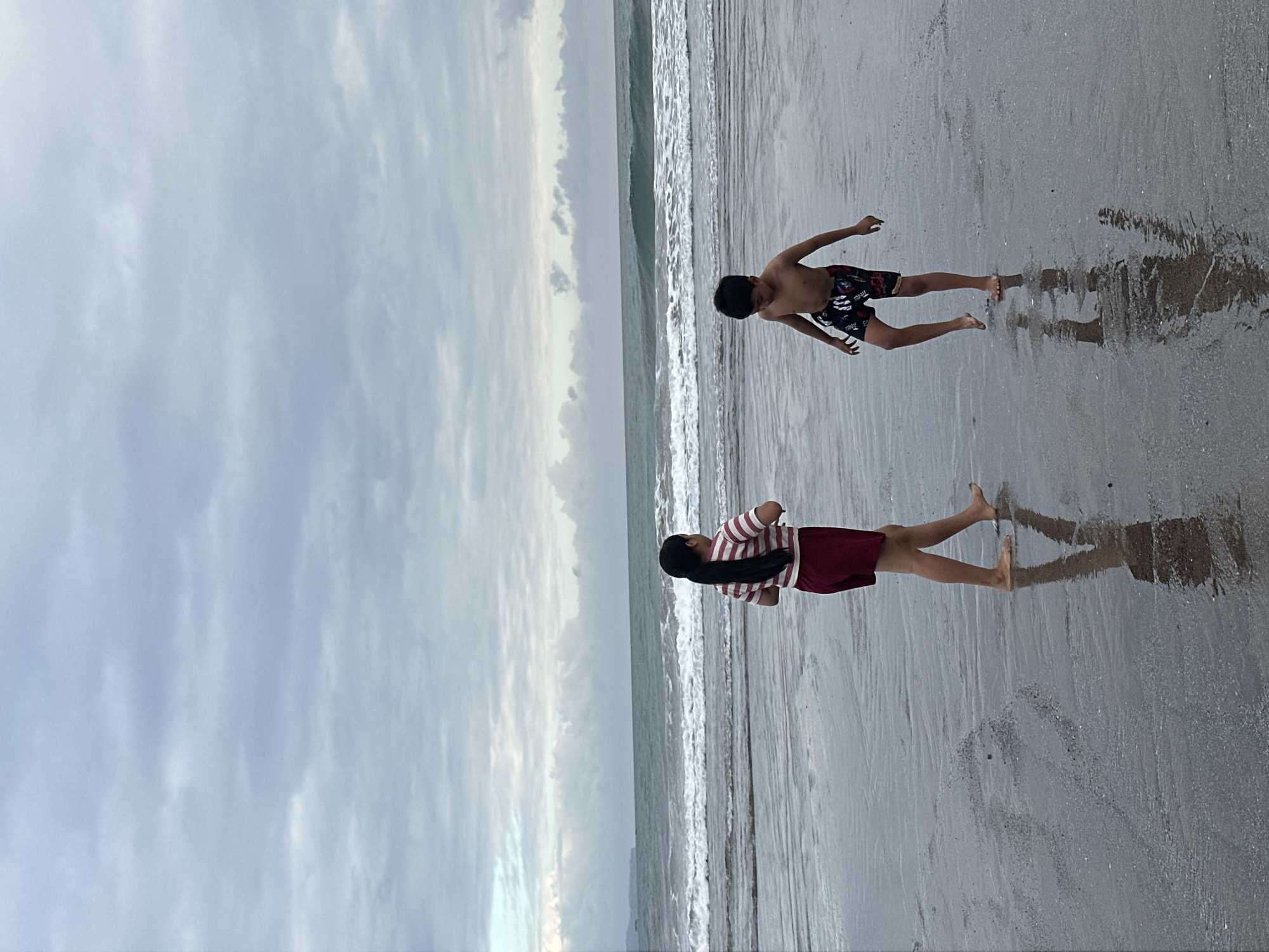 Two children, a girl and a boy, are standing on a wet sandy beach in front of a body of water. The girl, on the left, is wearing a striped red and white t-shirt and dark red shorts, and is facing away from the camera with her hands held together in front of her. The boy, on the right, is shirtless and wearing patterned black and red swim trunks, and is facing the girl with his arms extended outwards. The sand is wet and reflects the figures of the children. The ocean is visible behind them with moderate waves breaking on the shore. The sky is overcast with layers of grey and white clouds. The overall impression is a cool, grey day at the beach in North Kuta, Indonesia.