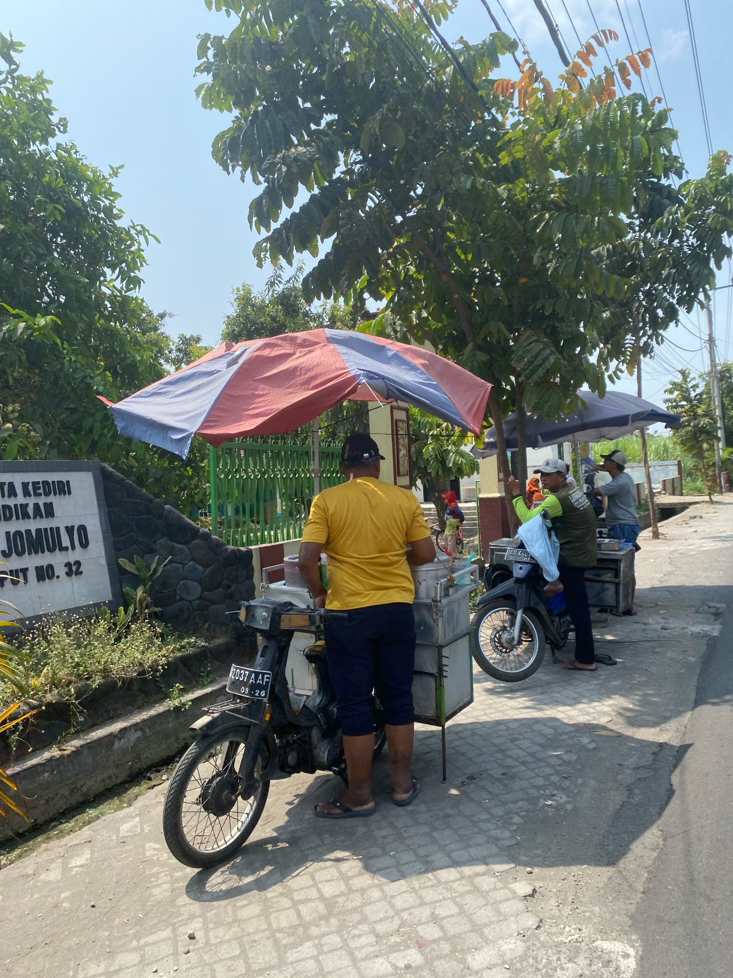 A street scene in Kediri City, Indonesia, shows two men selling food from mobile carts attached to motorcycles under large umbrellas. The man on the left, wearing a yellow shirt and dark pants, stands behind his cart, which is connected to a motorcycle with license plate 2037 AAF. The man on the right, wearing a green vest and dark pants, is engaged in a transaction with another person. Another man in a grey shirt is visible in the background. A stone wall with a sign that reads "TA KEDIRI DIKAN JOMULYO UT NO. 32" is to the left of the scene. Lush green trees and a clear blue sky form the background. The ground is paved with textured tiles and asphalt.