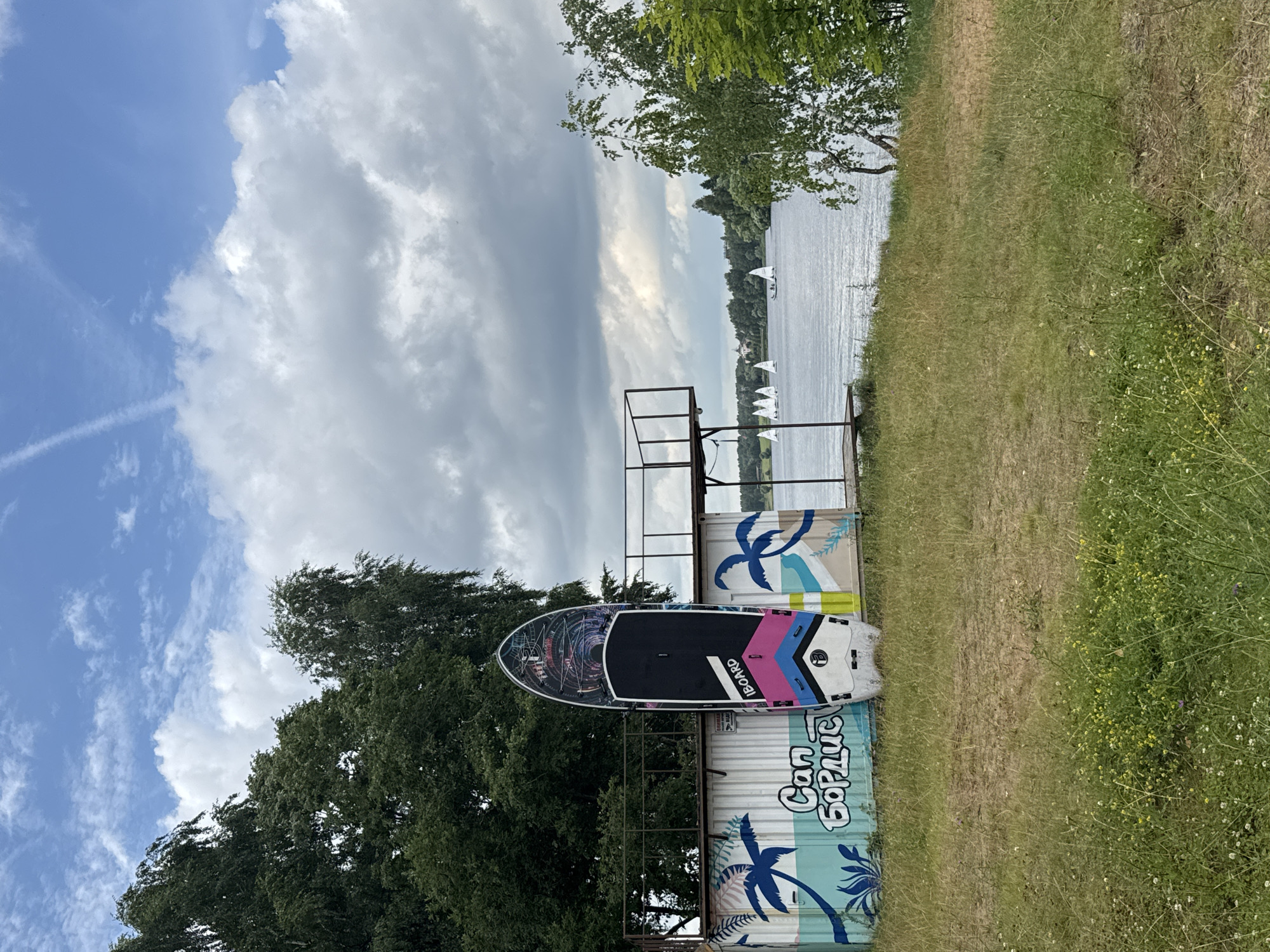 The image depicts a waterfront scene in Chiveryovo, Russia, under a partly cloudy sky. In the foreground, a grassy embankment slopes down towards a body of water. A stand-up paddleboard, decorated with abstract colorful designs and the "IBOARD" logo, is propped upright against a white, graffiti-adorned wall. The graffiti features stylized palm trees and the text "Can бордис". Behind the paddleboard and the wall, a metal platform structure with railings extends outwards.
The body of water, likely a lake or river, stretches across the mid-ground, reflecting the sky. Several small sailboats are visible on the water in the distance, along with a treeline and buildings on the opposite shore. Lush green trees frame the scene, with one prominent birch tree with white bark on the right side of the image. The overall atmosphere is that of a recreational area by the water.