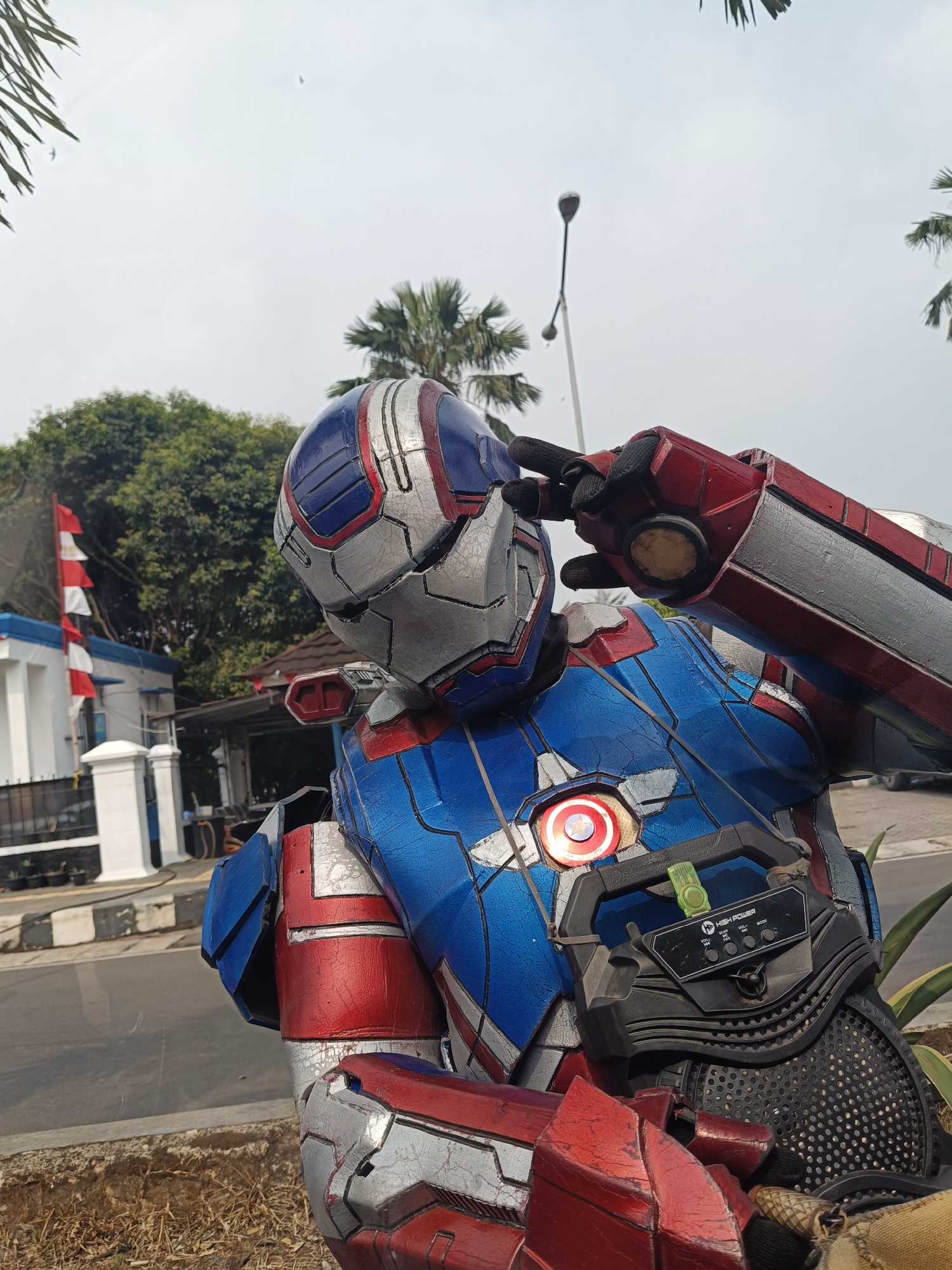 A person wearing a red, white, and blue armored suit, resembling the Iron Patriot from Marvel comics, is shown from the chest up. The suit has a metallic finish with visible paint chipping, giving it a weathered appearance. A red and blue striped helmet covers the individual's head, with a hand raised to their forehead in a salute. A circular emblem resembling Captain America's shield is visible on the chest plate, emitting a soft red light. Attached to the suit's chest is a black portable speaker with various buttons and labels. The figure is positioned outdoors, with palm trees and other foliage in the background. To the left, a white building with blue trim and a red and white Indonesian flag on a flagpole are visible. A street with a black and white striped curb runs in front of the building. The location is identified as the city of Pandeglang, Indonesia.