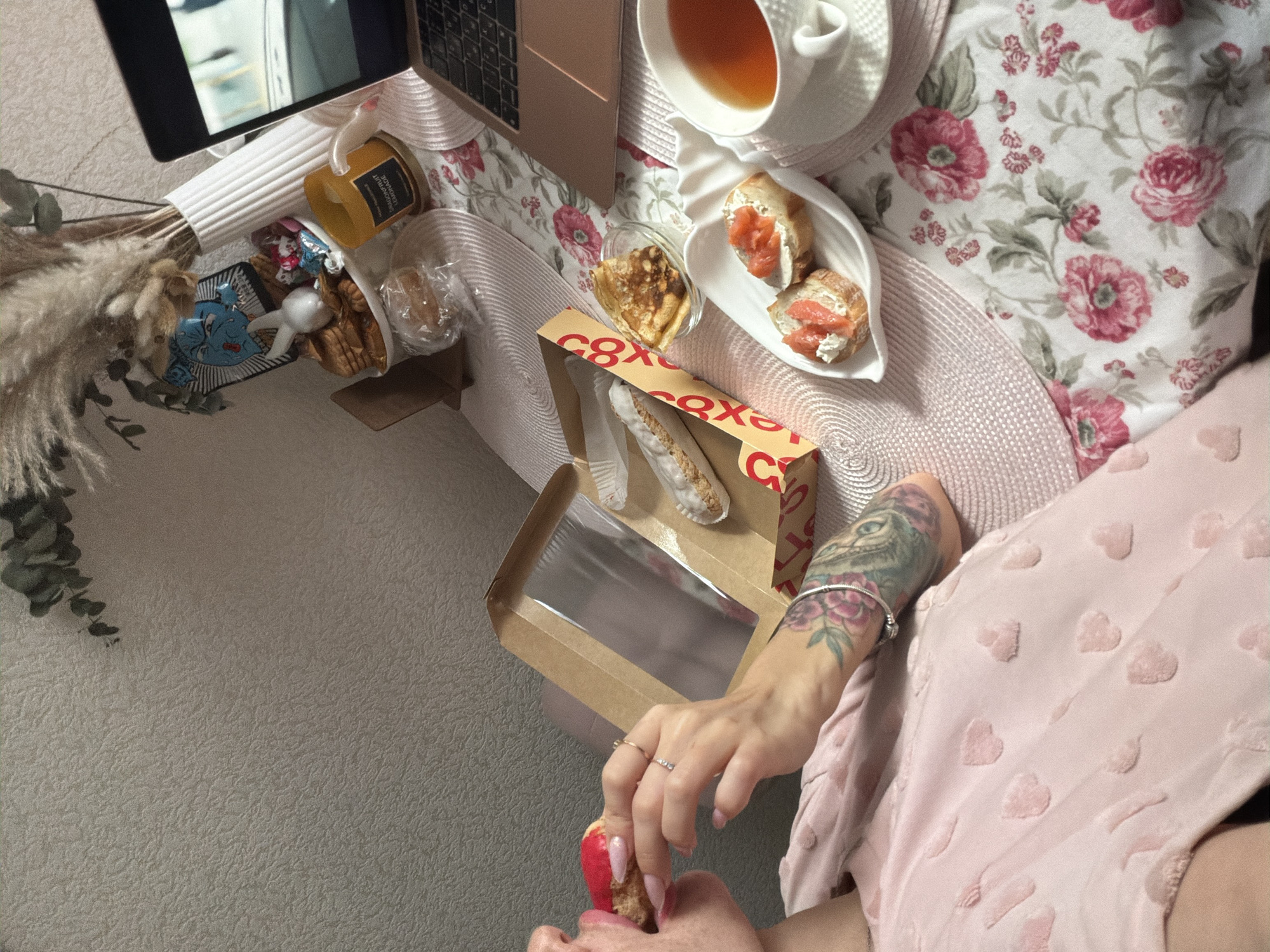 The image depicts a person sitting in what appears to be an indoor setting. The person has a tattooed arm and is wearing a light pink top with heart patterns. They are holding and eating a brightly colored pastry, possibly a macron.
On the surface next to the person are several items: a laptop, a cup of tea, a plate with rolls topped with salmon, a small dish of what looks like crepes, and a box of pastries. Other items include decorative plants, a decorative candle, a container with candy and a small ceramic figure.
The image does not provide enough information to definitively identify the location as Saint Petersburg, Russia. It is a personal, domestic scene.