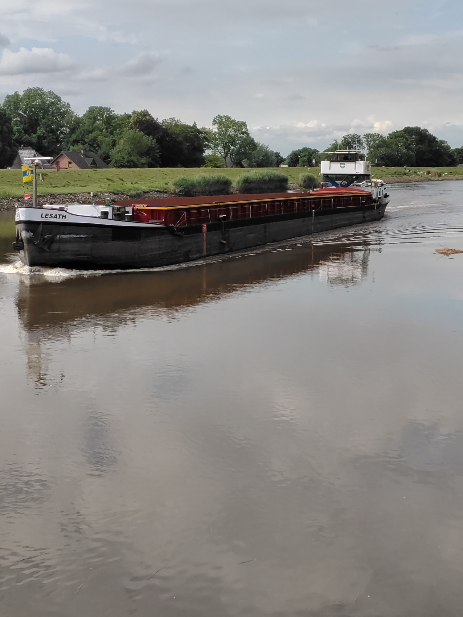 The image shows a barge named "LESATH" traveling on a river in Heiligenstedten, Germany. The barge is dark gray with a red cargo cover and a white superstructure at the stern. The name "LESATH" is visible in black lettering on the bow of the barge. The barge is creating a wake as it moves through the water. On the left bank of the river, there are houses and trees. The sky is overcast with some clouds. The river water is a muddy brown color and reflects the sky and the barge. The overall scene depicts a working barge navigating a waterway in a rural or semi-rural German setting.