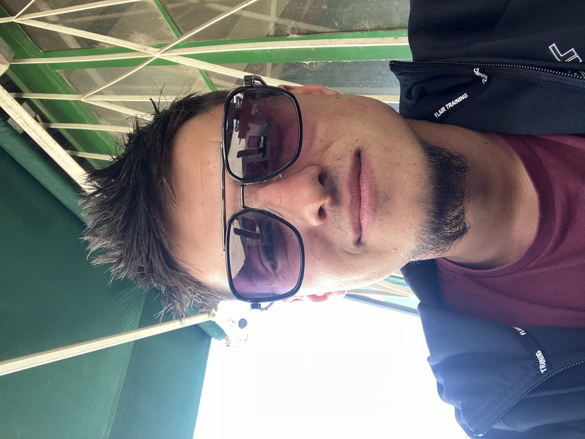 A man with dark hair and a beard is wearing sunglasses and a burgundy shirt underneath a black jacket. "FLAIR TRAINING" is printed on the jacket. He is likely sitting under a structure with a green awning and a patterned ceiling. The image does not give any information about the location in Krastevich, Bulgaria.