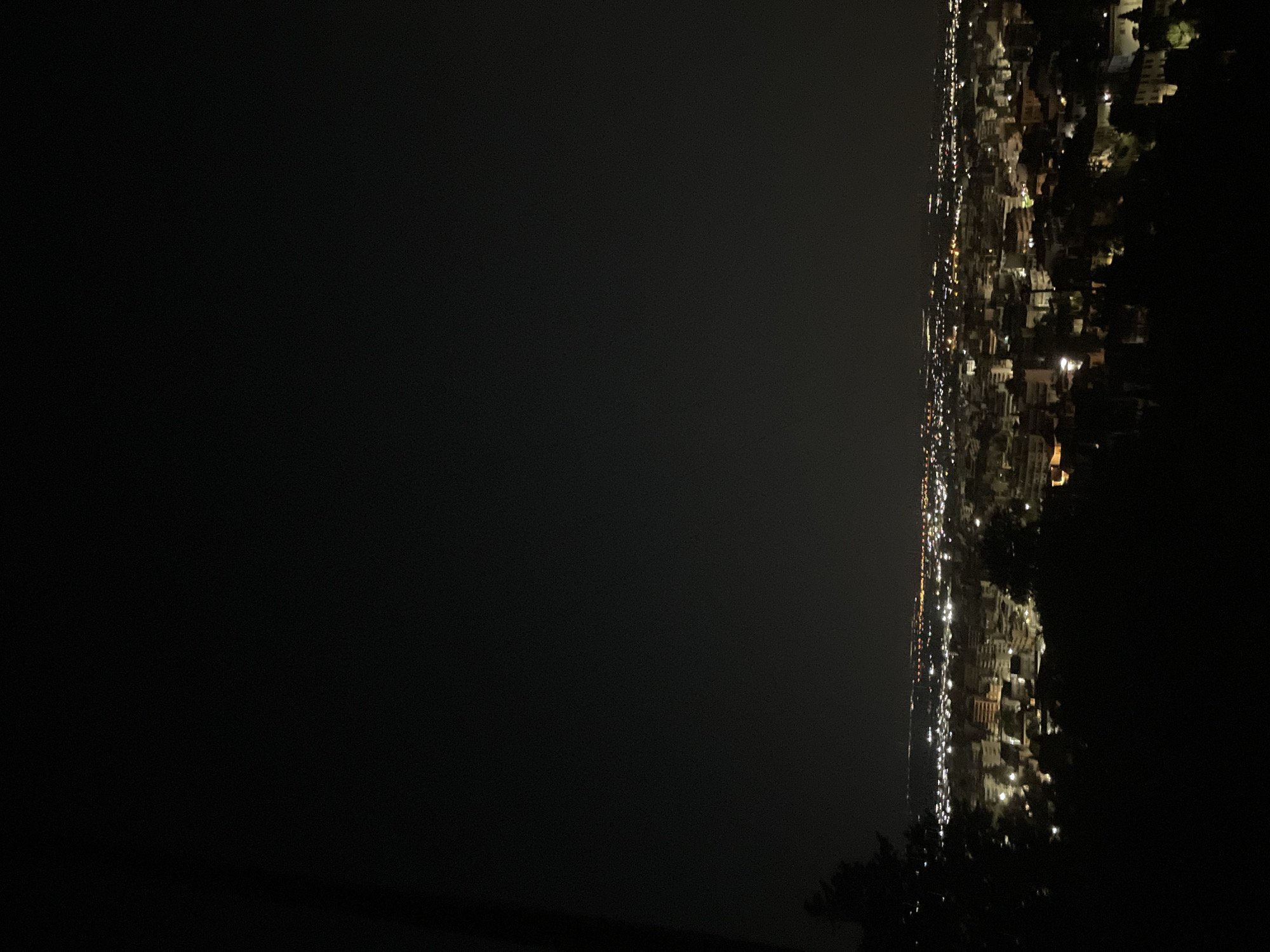 An aerial night view of the city of Xanthi, Greece, is captured from a hillside overlooking the urban landscape. The city lights create a warm, diffused glow against the dark sky, illuminating the dense network of buildings. The foreground is dominated by the dark silhouettes of trees and foliage, framing the view of the city below. The perspective suggests the photograph was taken from a vantage point at a significant elevation. The lights are a mix of yellow and white, indicating streetlights and illuminated windows. The overall impression is a serene and sprawling urban scene at night.