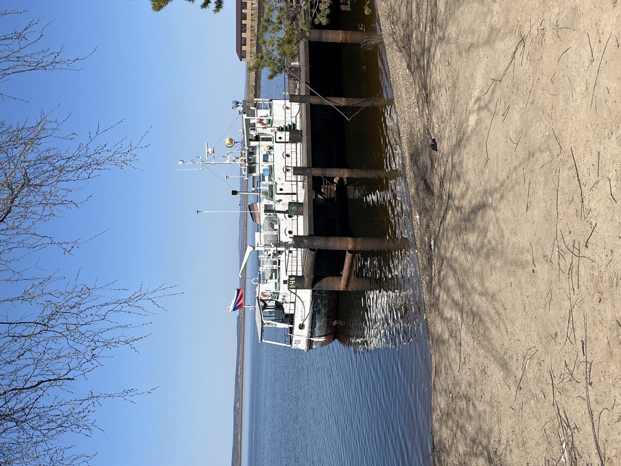 The image depicts a docked ship, identified by the name "Я-4646" and a Russian flag, at what appears to be a pier or dock. The location is likely in Chaikovsky, Russia, given the presence of the Russian flag and Cyrillic script on the ship. The ship is white with dark trim and has multiple levels. The water is calm with reflections of the ship and pier. The pier or dock is made of concrete and there is some dirt and vegetation visible on the adjacent land. A bare tree is to the left of the image and a building is to the right of the image. The sky is clear and blue.