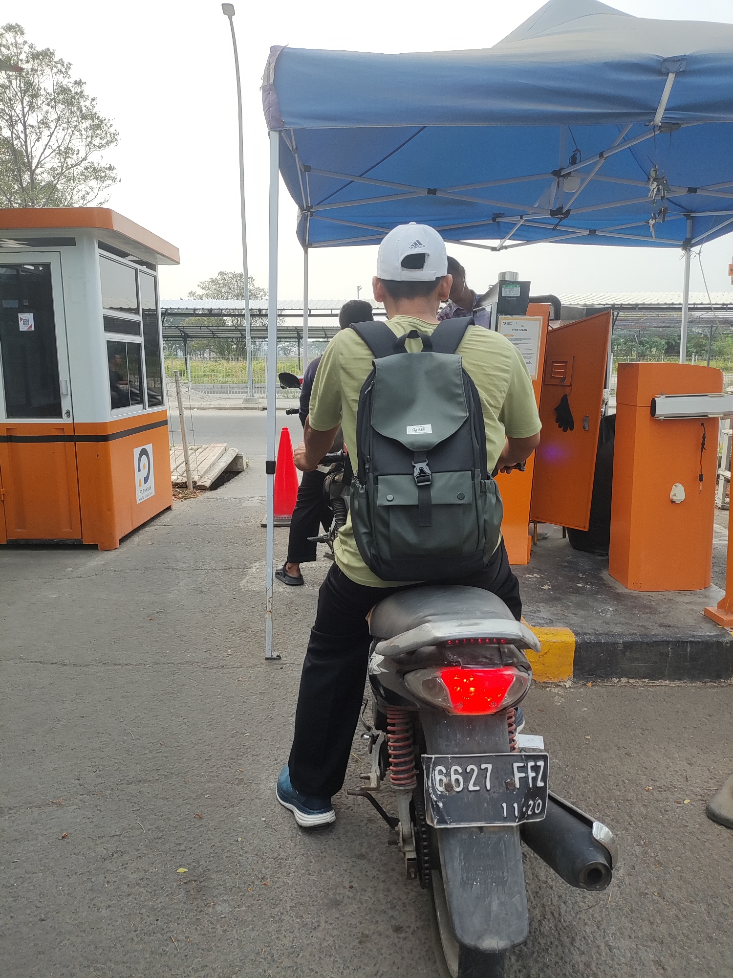 A person wearing a green t-shirt, black pants, and a white Adidas baseball cap is sitting on a motorcycle at what appears to be a toll booth. The person is wearing a dark green backpack. The motorcycle has a license plate that reads "6627 FFZ" and "11/20". The background shows an orange toll booth structure, a blue canopy tent, and some trees. The ground is paved asphalt. The scene appears to be outdoors in daylight.