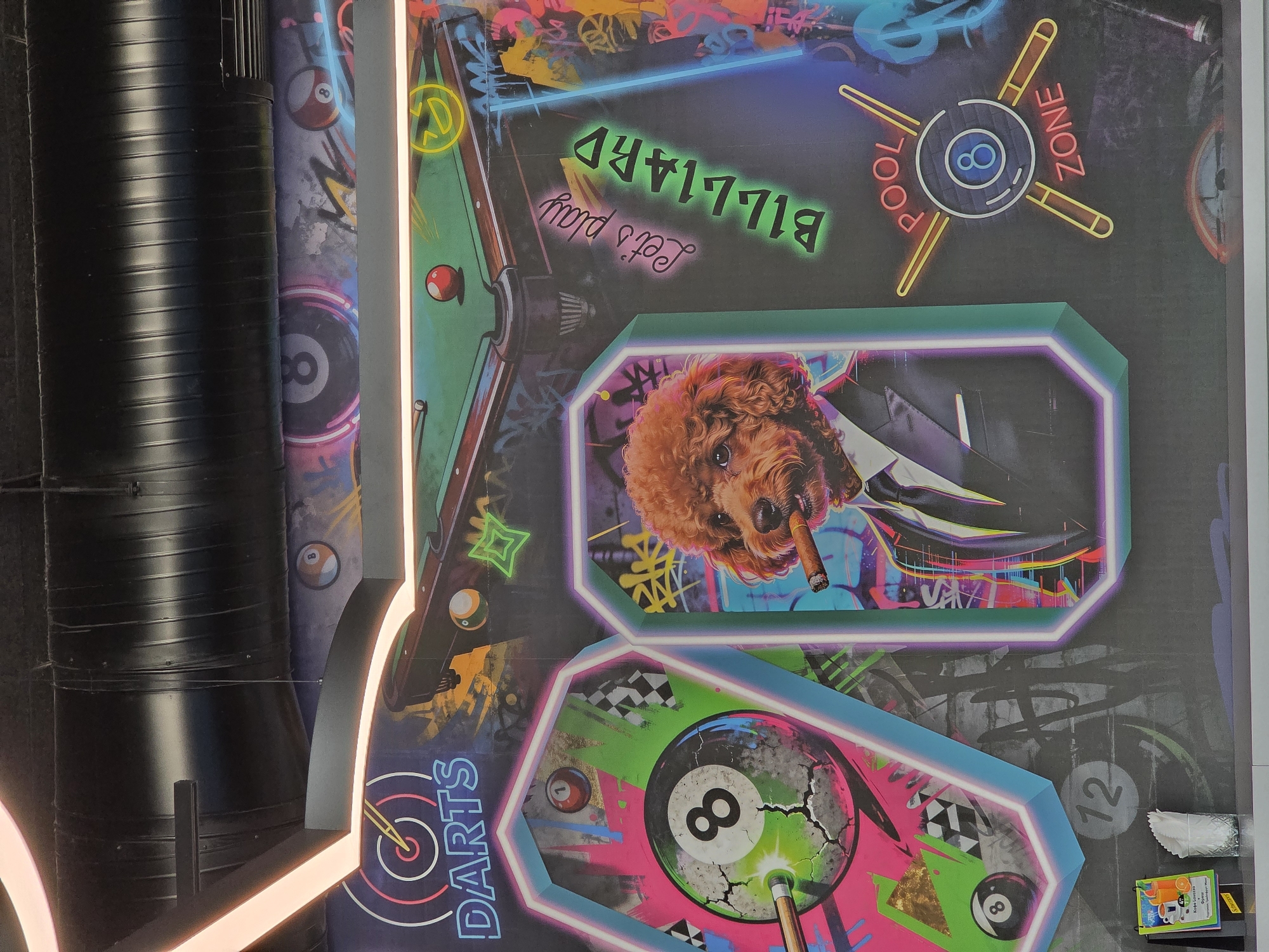 The image depicts an indoor wall mural with neon-style designs, likely in a commercial or entertainment space. The designs feature a pool table, a dog in a suit smoking a cigar, darts, and an eight-ball, all with bold, bright colors. The mural also incorporates the phrases "Let's play Billiard," "Pool Zone," and "Darts." The environment includes visible ventilation pipes and menu books and napkins on the wall. Varna, Bulgaria, is mentioned only as a possible location, and no specific details confirm this placement. There are no people in the image.