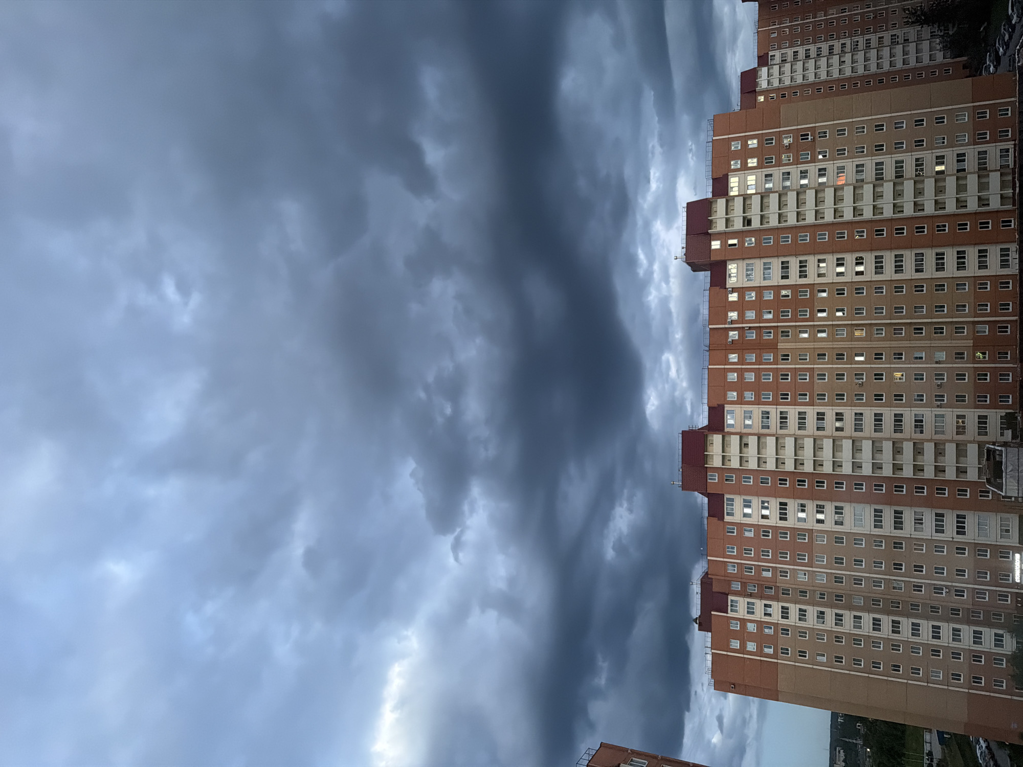 A medium shot captures a multi-story apartment building against a dramatic, overcast sky. The building, which appears to be constructed of brick and concrete, features numerous windows, some of which are illuminated, suggesting occupied apartments. The sky is filled with dark, heavy clouds, indicating impending bad weather. The scene is framed from a slightly elevated perspective, looking down upon the building. The overall mood is somber, characterized by the dark clouds and the muted tones of the building. The image is set in an urban environment, with hints of distant trees and parked cars visible at the base of the building. The location is not specified.