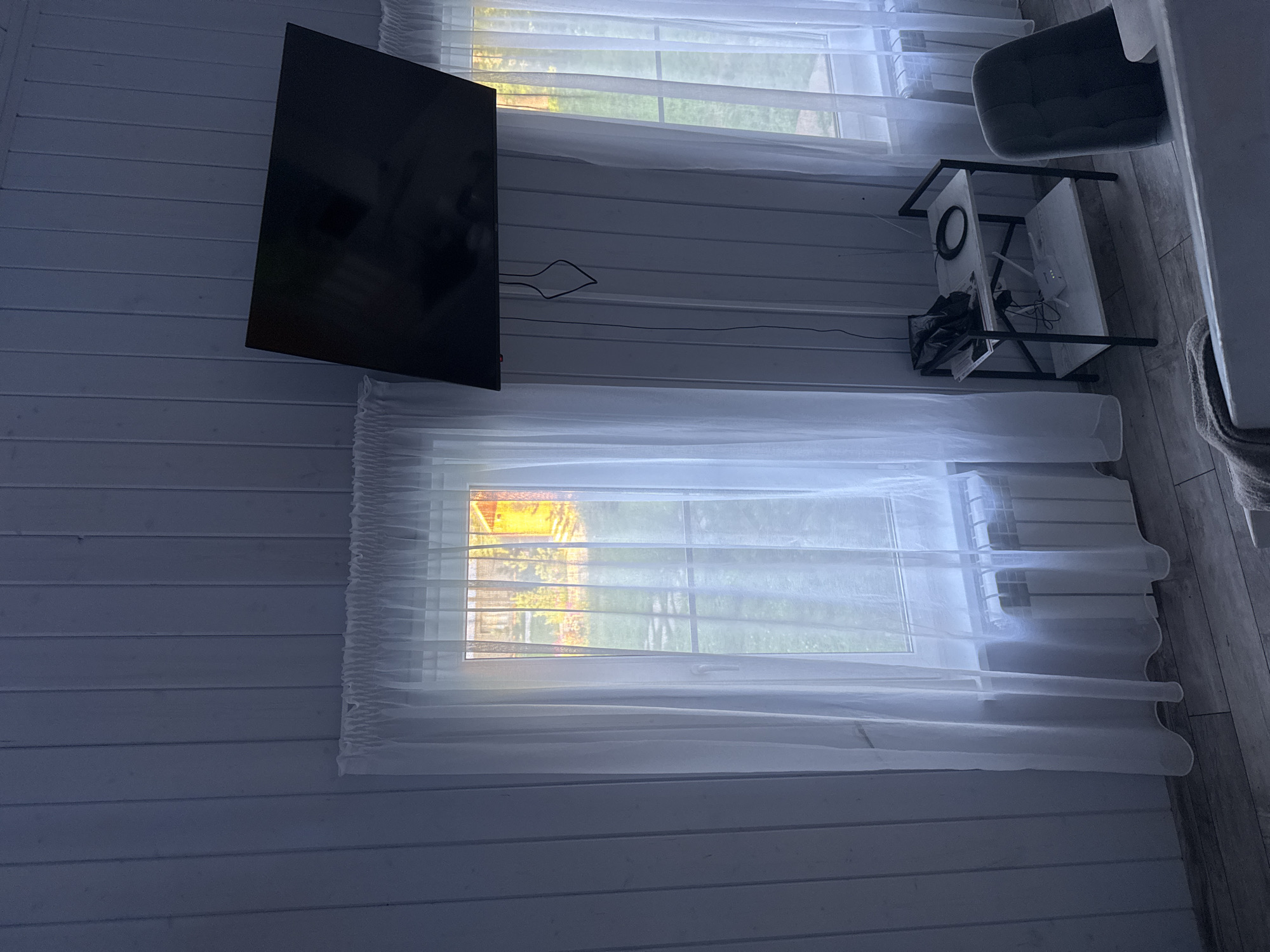 The image depicts an interior scene, likely a room in a building located at Doma 1096 Km, Russia. The walls are paneled with light-colored vertical planks. Two windows are visible, each covered with sheer white curtains. Through the left window, a sunlit yard with greenery and a structure is visible, illuminated by warm, golden light. The right window shows a similar outdoor scene, though less distinct.
Mounted on the wall above the windows is a large, flat-screen television, angled downwards. Black cables hang from the television, extending towards a small, two-tiered shelving unit positioned to the right of the left window and below the television. On the top shelf of the unit are some items, including what appears to be a black plastic bag and a coiled cable. On the bottom shelf, a white wireless router and some other electronic devices are present.
To the right of the shelving unit is a dark gray upholstered chair. The floor is made of light-colored wooden planks. A white radiator is situated beneath the left window. The overall lighting in the room is dim, with the primary light source appearing to be from the windows. No people are present in the image.