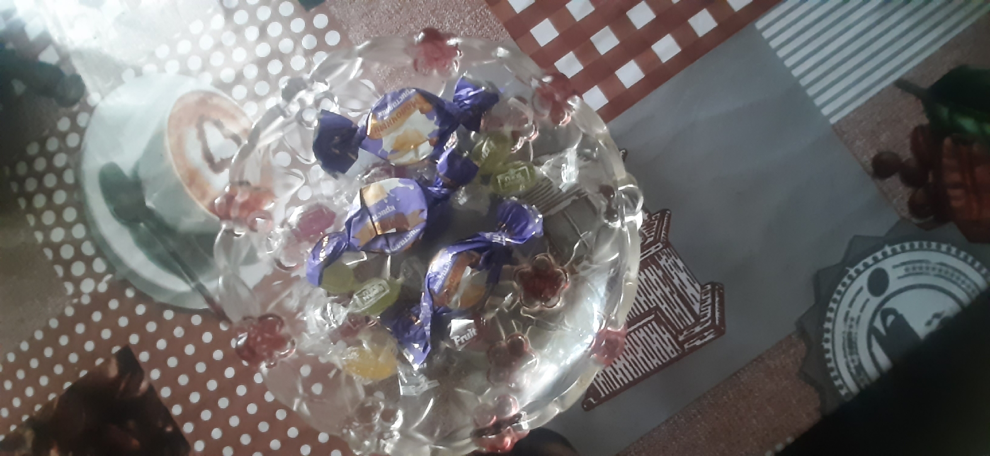 The image shows a glass bowl filled with individually wrapped candies. The candies are wrapped in purple, gold, and light green foil. Some of the wrappers have text visible, including "Fruit" and what appears to be Russian text. The bowl itself is clear glass with decorative floral elements along the rim. It is placed on a table with a brown and white polka dot patterned tablecloth. In the background, there is a white plate with a dark substance, possibly coffee or chocolate, and a spoon. The scene is likely indoors, given the lighting and the presence of a tablecloth. There are no people visible in the image. The location of Yessentukskaya, Russia is not depicted or suggested by any elements within the image itself.
