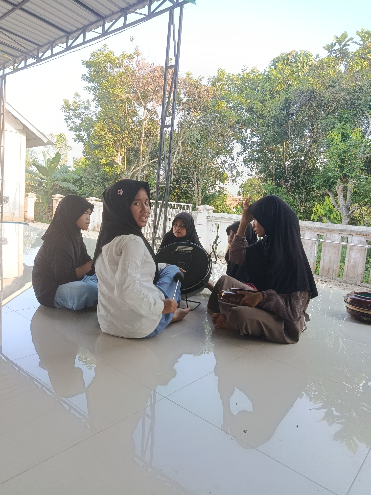 Four young females, wearing hijabs and long-sleeved clothing, are sitting on a tiled outdoor floor. The individual in the foreground, facing left, is wearing a white shirt and blue jeans. To her left, another female in a black hijab and black shirt sits facing away from the camera. Between these two and further back, a third female in a black hijab is partially visible. To the right, a fourth female in a black hijab and brown patterned clothing is gesturing with her hands while holding a small instrument in her lap. A large drum is situated in front of the female in the white shirt, and another smaller drum is to the far right. The setting appears to be a covered outdoor patio with a white railing and lush green trees in the background, suggesting a residential area in Swarga Bara, Indonesia. The floor is reflective, showing the reflections of the individuals and the surrounding foliage.