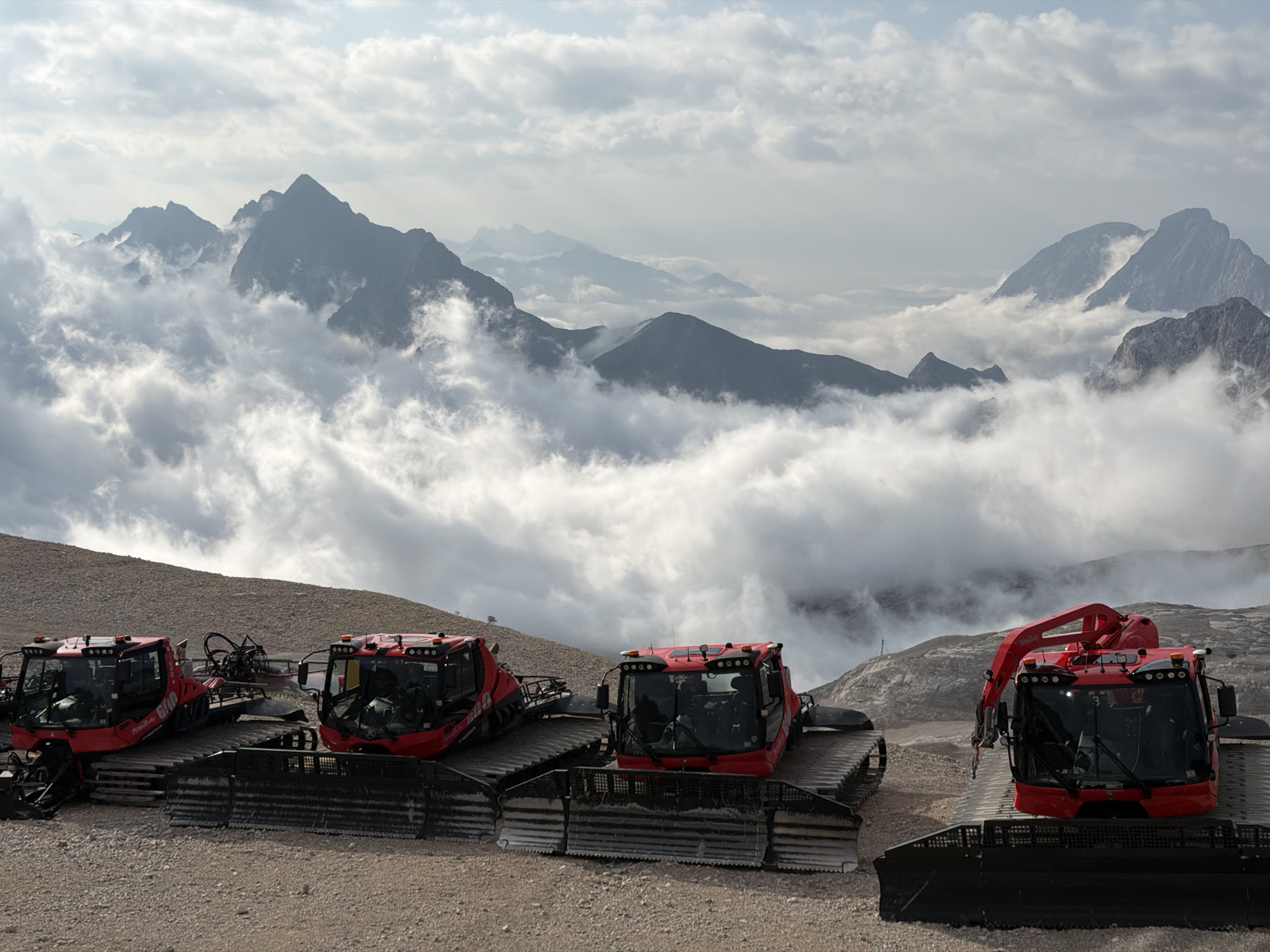 The image shows a mountain landscape with four red snow groomers parked on a gravelly surface. In the background, majestic mountains are visible through a layer of thick clouds. The sky is overcast with some areas of sunlight breaking through. The snow groomers, also known as Pistenbullys, are equipped with tracks and large blades at the front, indicating their use in maintaining ski slopes or trails. The scene appears to be at a high altitude, likely on a mountain resort. While the specific location is not visually identifiable, Garmisch-Partenkirchen, Germany, is known for its mountainous terrain and ski resorts, making it a plausible general setting. There are no people visible in the image.