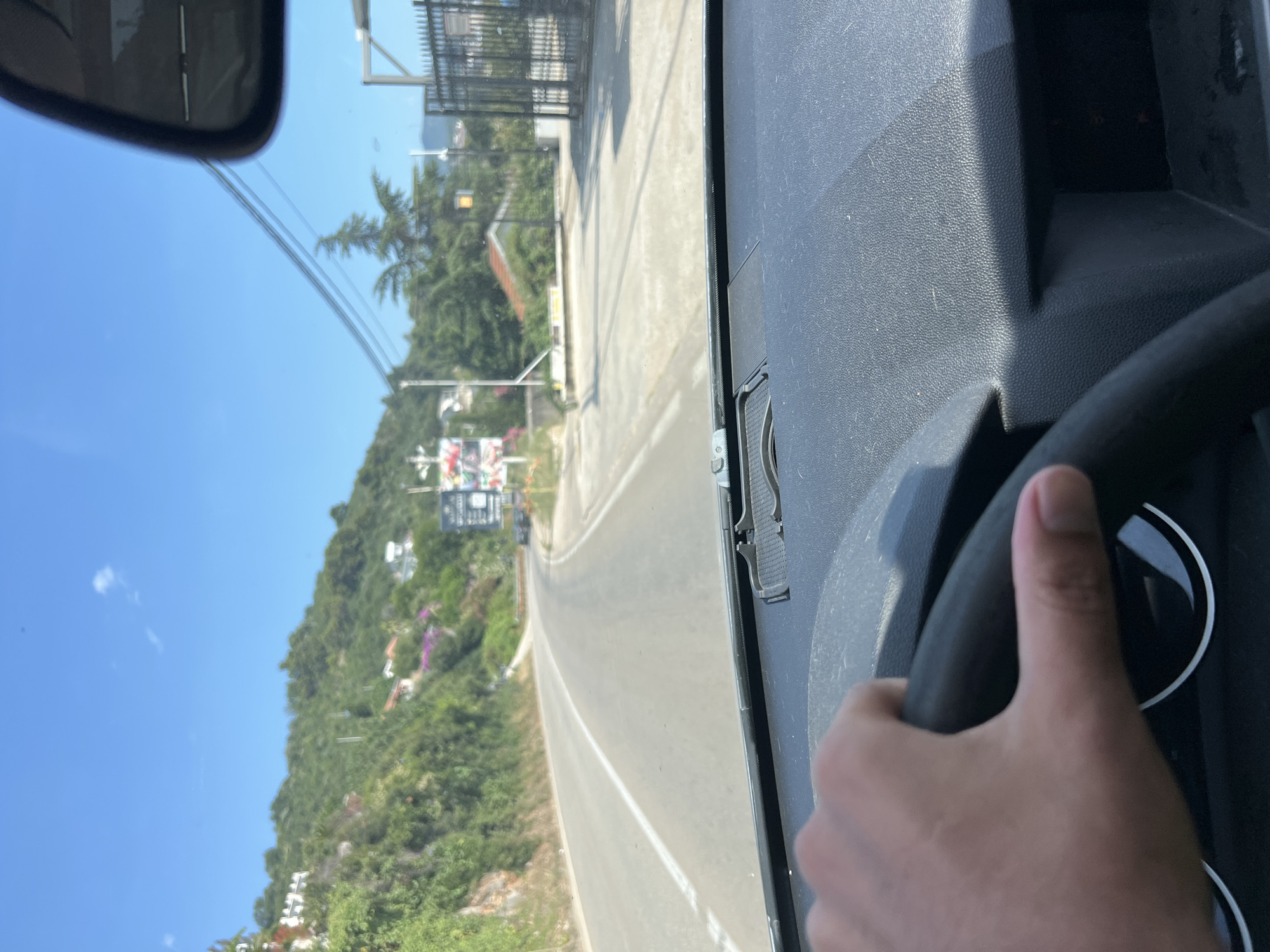 The image is taken from the interior of a vehicle, showing a hand gripping the steering wheel. The view is through the windshield, looking forward onto a paved road that curves to the left. The road is bordered by lush green vegetation on the left, with some white buildings visible on the hillside. On the right side of the road, there are trees, a dark metal fence, and some structures that appear to be commercial. In the distance, there are more hills and buildings. The sky is clear and blue with a few small white clouds. The scene appears to be a sunny day in a coastal or hilly region, likely Dobra Voda, Montenegro, given the presence of Mediterranean vegetation and architecture.