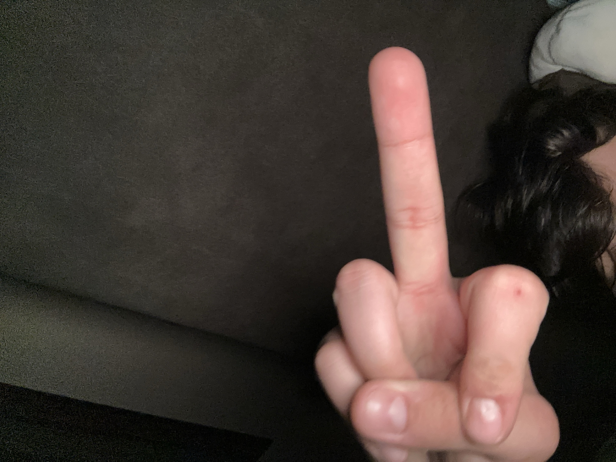 A person's hand is giving the middle finger. The person has dark curly hair and is lying down, possibly in bed. The background is dark. The location is unknown.