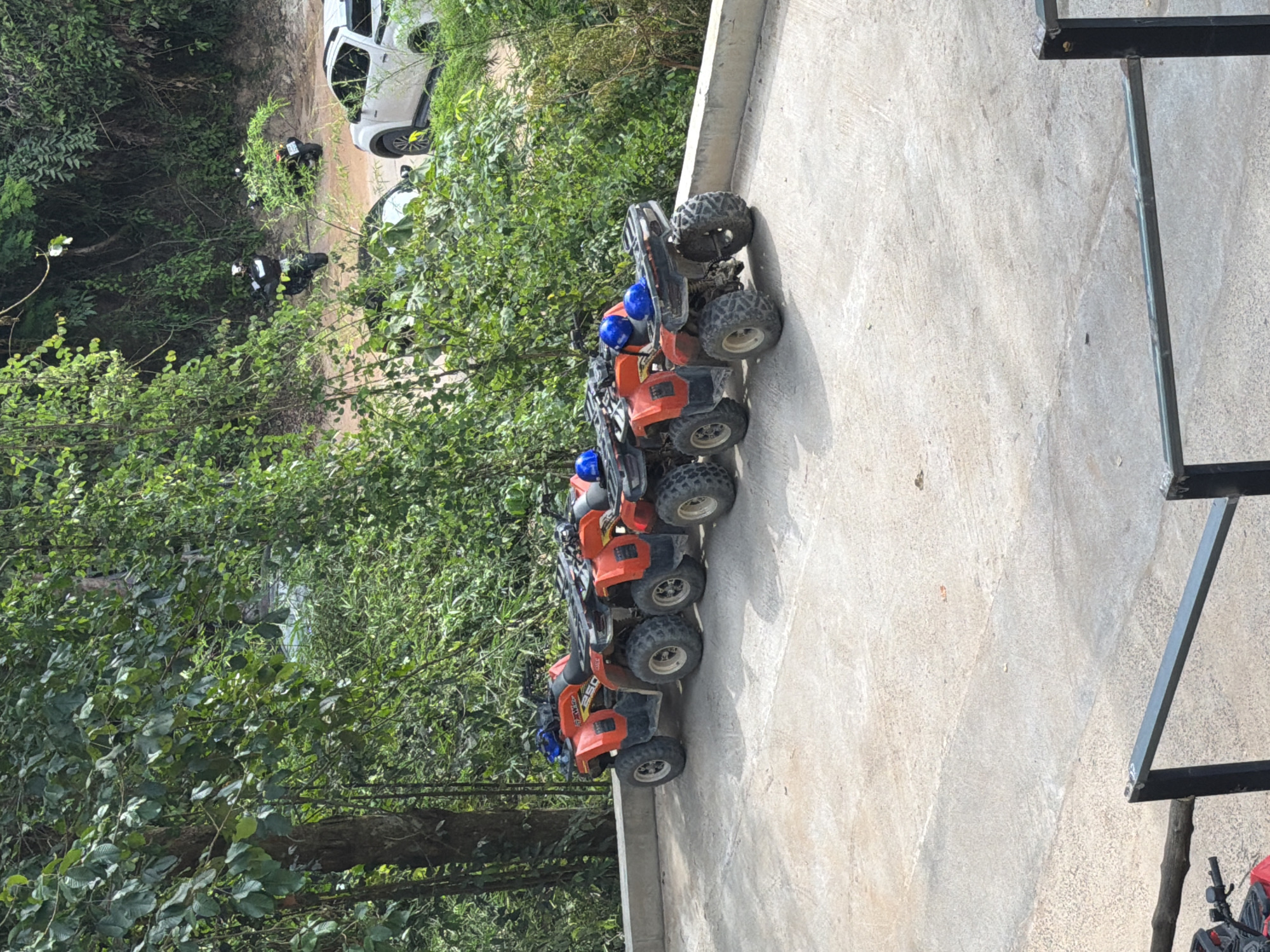 In Ko Samui, Thailand, a concrete parking area is lined with four red all-terrain vehicles (ATVs) on the left side of the image, each equipped with blue helmets. The ATVs are parked parallel to a low concrete wall that separates them from a lush, green, tree-covered hillside. In the background, a dirt road winds up the hill, where two individuals on motorcycles are visible, one wearing a black and white outfit and the other in a dark outfit. A white pickup truck is also parked further up the road. The environment suggests an outdoor adventure or recreational area, likely a starting point for ATV or motorcycle tours within the tropical landscape of Ko Samui.