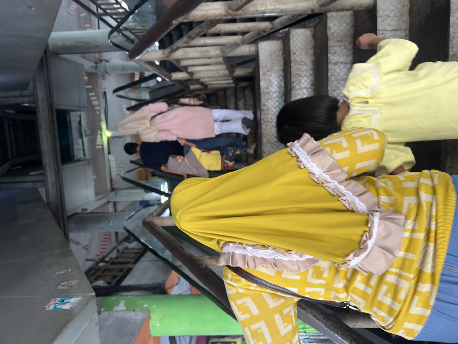 A stairwell ascends amidst a family. Several individuals are visible on different stair levels. One person at the front wears a yellow dress and hijab with a distinctive pattern, followed by a child in yellow clothing. Behind them, other family members are dressed in casual attire, including a pink sweatshirt, blue jeans, and a navy blue jacket. The stairwell features metal stairs and handrails, with architectural details suggesting an aged or industrial building.