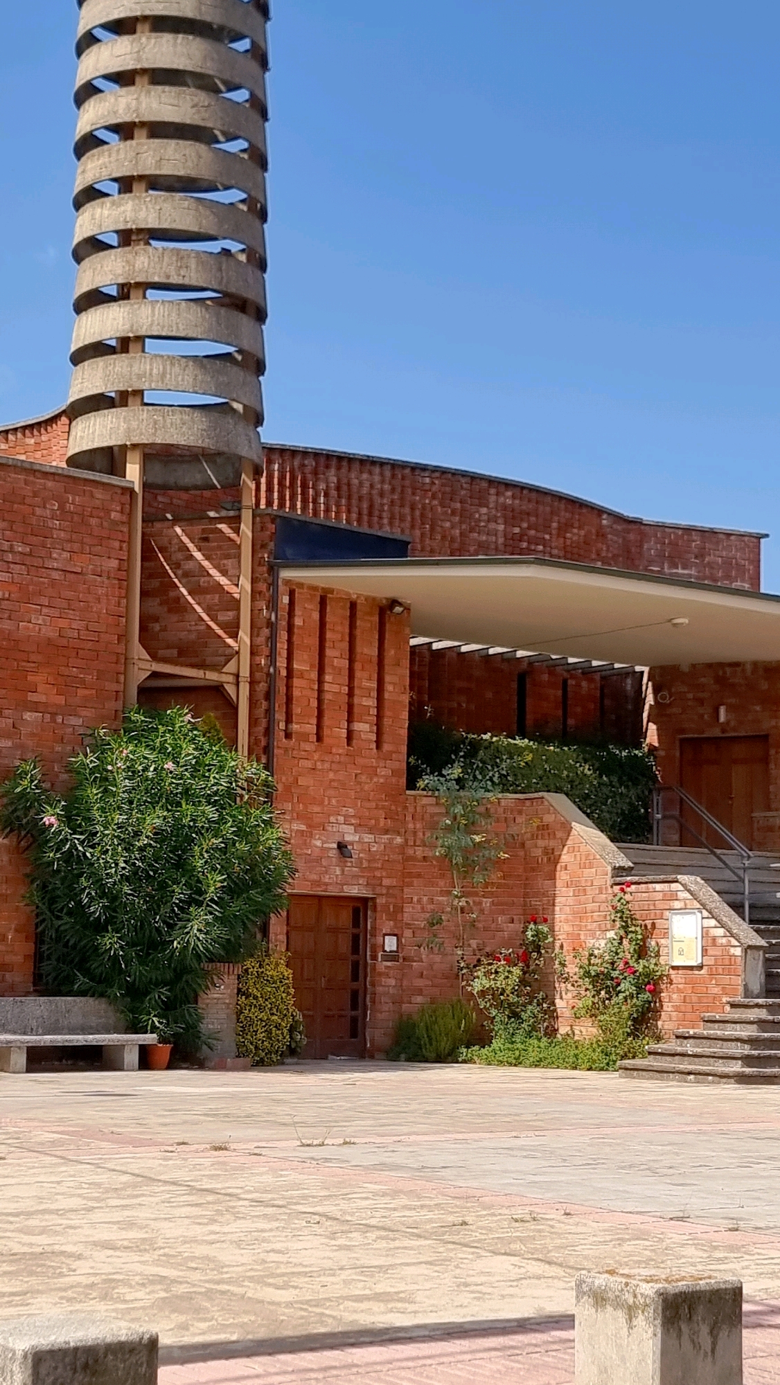 The image depicts the exterior of a modern building constructed primarily of red brick. A prominent cylindrical structure with horizontal concrete rings encircles a wooden frame, extending vertically upwards. Adjacent to this structure, the building features a series of vertical indentations on its brick facade. A covered walkway with a white ceiling and exposed supporting beams extends from the building. Green foliage, including a large bushy plant and climbing roses with red blooms, is visible in front of the brick walls and around a set of outdoor stairs. A concrete bench is situated to the left, near a wooden door. The ground is paved with concrete, featuring sections of contrasting colors. The scene is set against a clear blue sky. The building is located in La Canya, Spain. There are no people visible in the image.