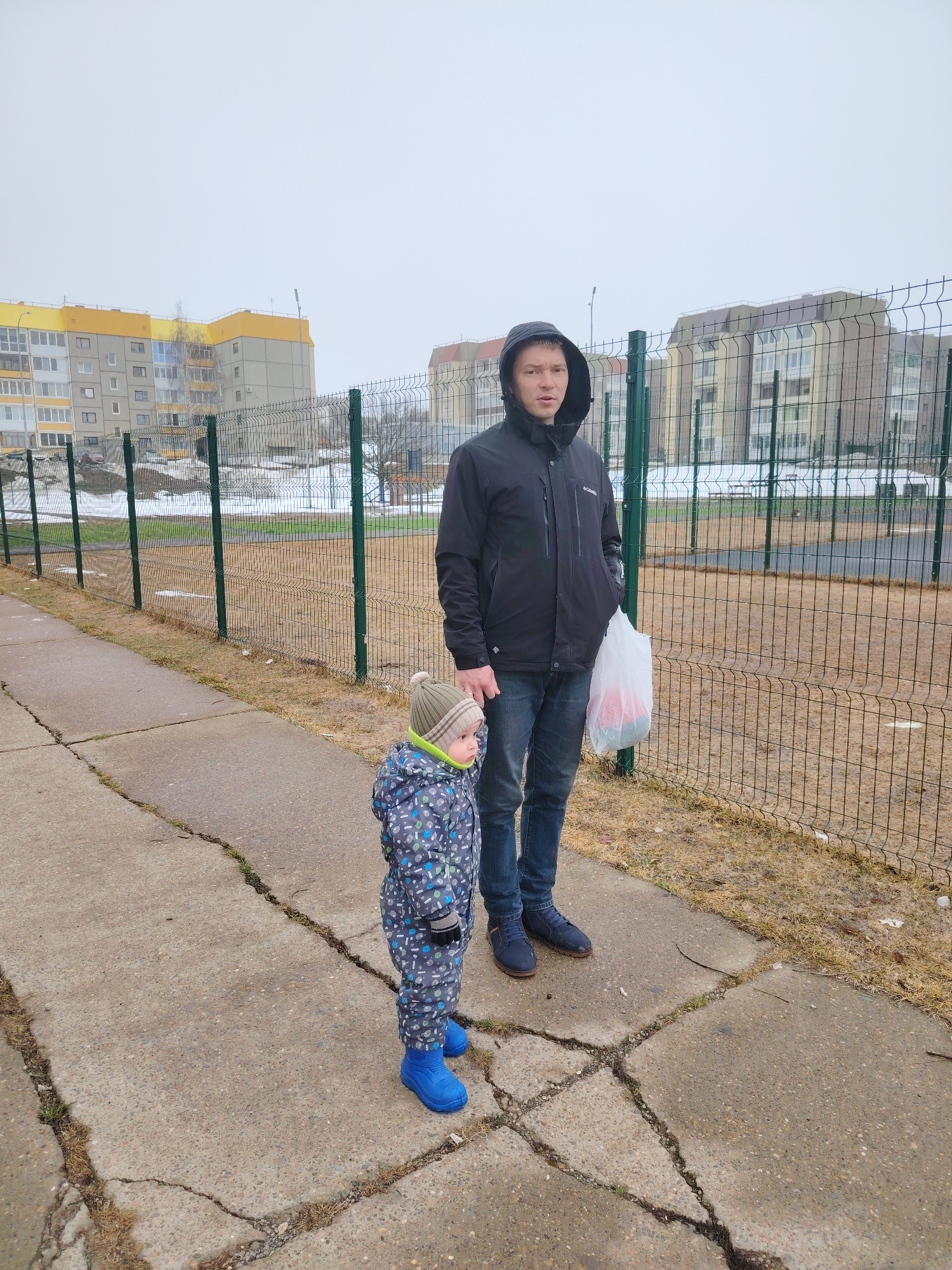 A man and a small child are standing on a cracked concrete sidewalk next to a green wire fence. The man is wearing a black hooded jacket, blue jeans, and blue shoes. He is holding a white plastic bag. The child is wearing a patterned snowsuit, a knitted hat, and blue boots. In the background, there are several apartment buildings and a playing field. The location is Марковский, Russia. The day is overcast. Patches of snow are visible on the ground.
