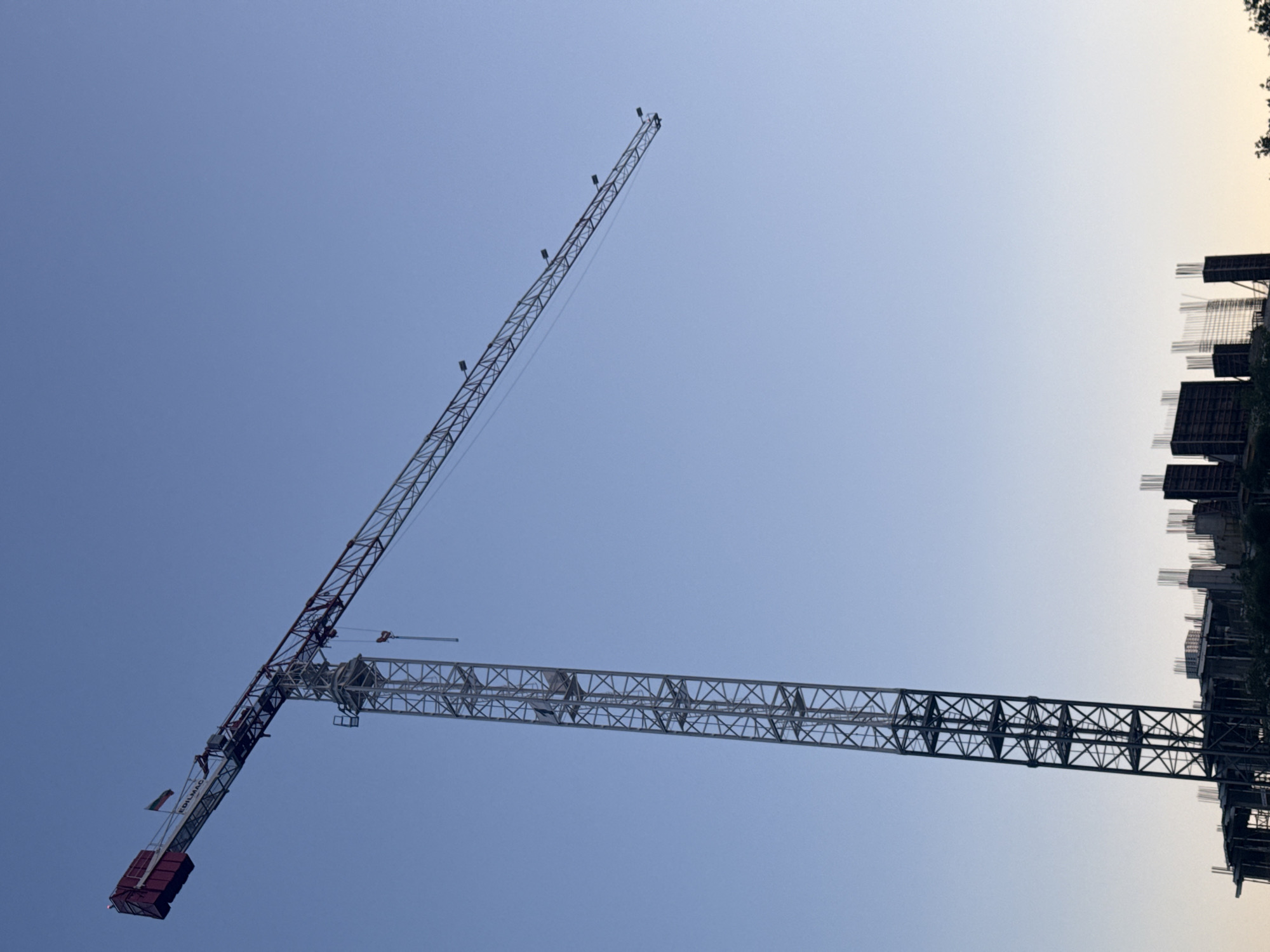 A tall, grey construction crane with a red cab and boom extends diagonally across a clear, pale blue sky. The crane's boom stretches horizontally to the right, with a hook and cable hanging from it. Below the crane, a partially constructed building with exposed rebar is visible. The overall scene appears to be a construction site in the city of Burgas, Bulgaria.