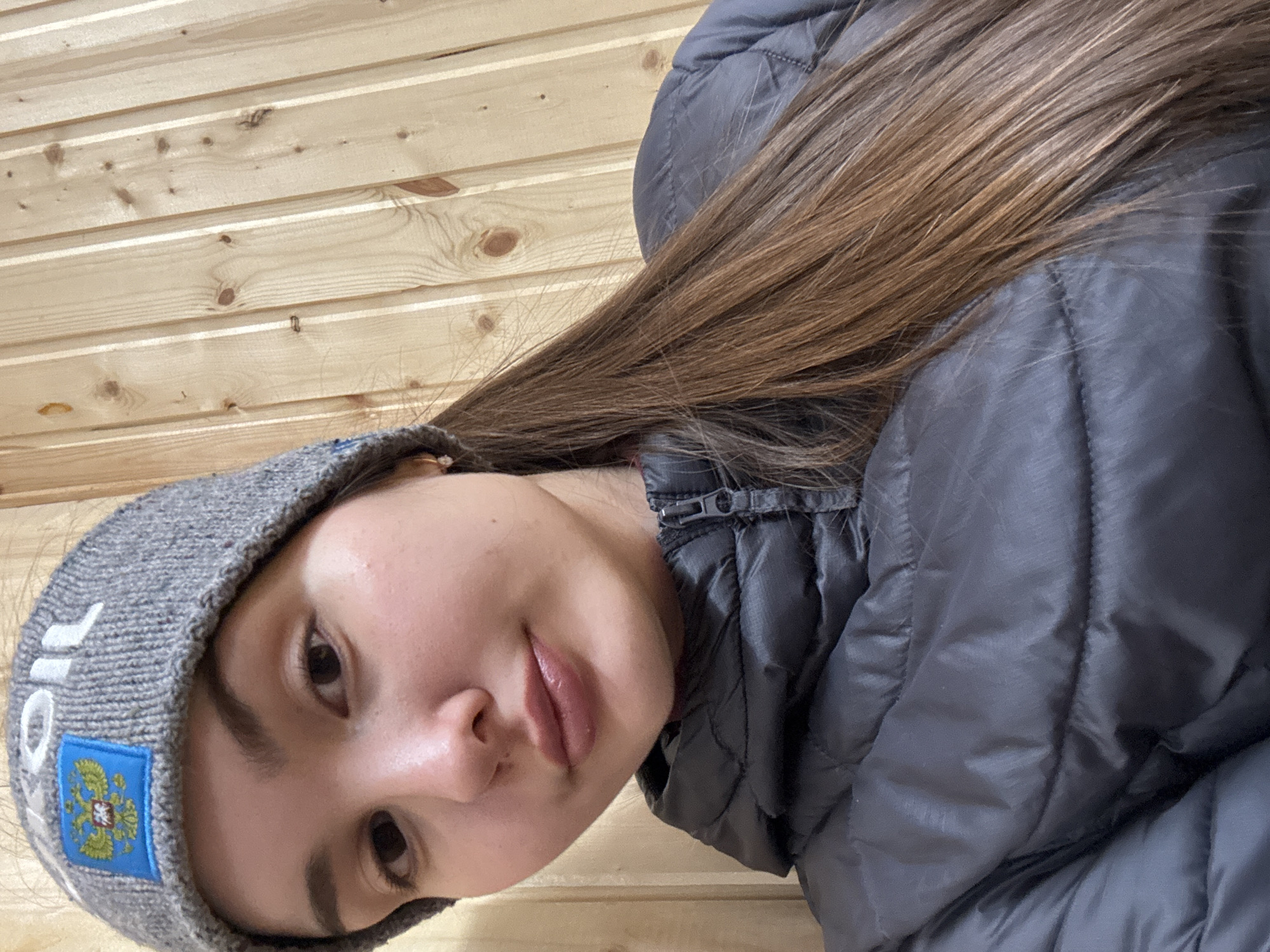 A woman is in front of a light wood wall. The woman has long brown hair and is wearing a gray beanie with a patch on it. The patch on the beanie has an image of a heraldic double eagle with a red shield. She is also wearing a gray puffy jacket. The city is Orenburg, Russia.