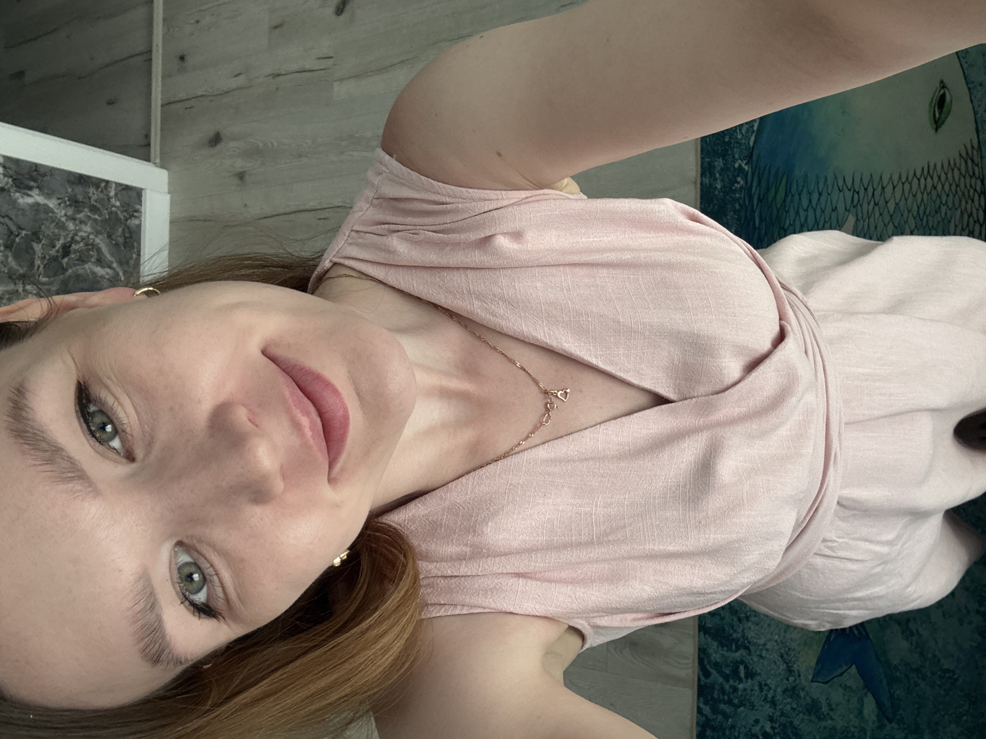 A woman with light skin, light green eyes, and light brown hair is facing the camera. She is wearing a light pink dress and a gold necklace with heart pendants. She is indoors. The background includes a light wood-patterned floor, a countertop with marble, and a blue rug with a fish pattern. The location is not identifiable from the image alone as being in Izhevsk, Russia.