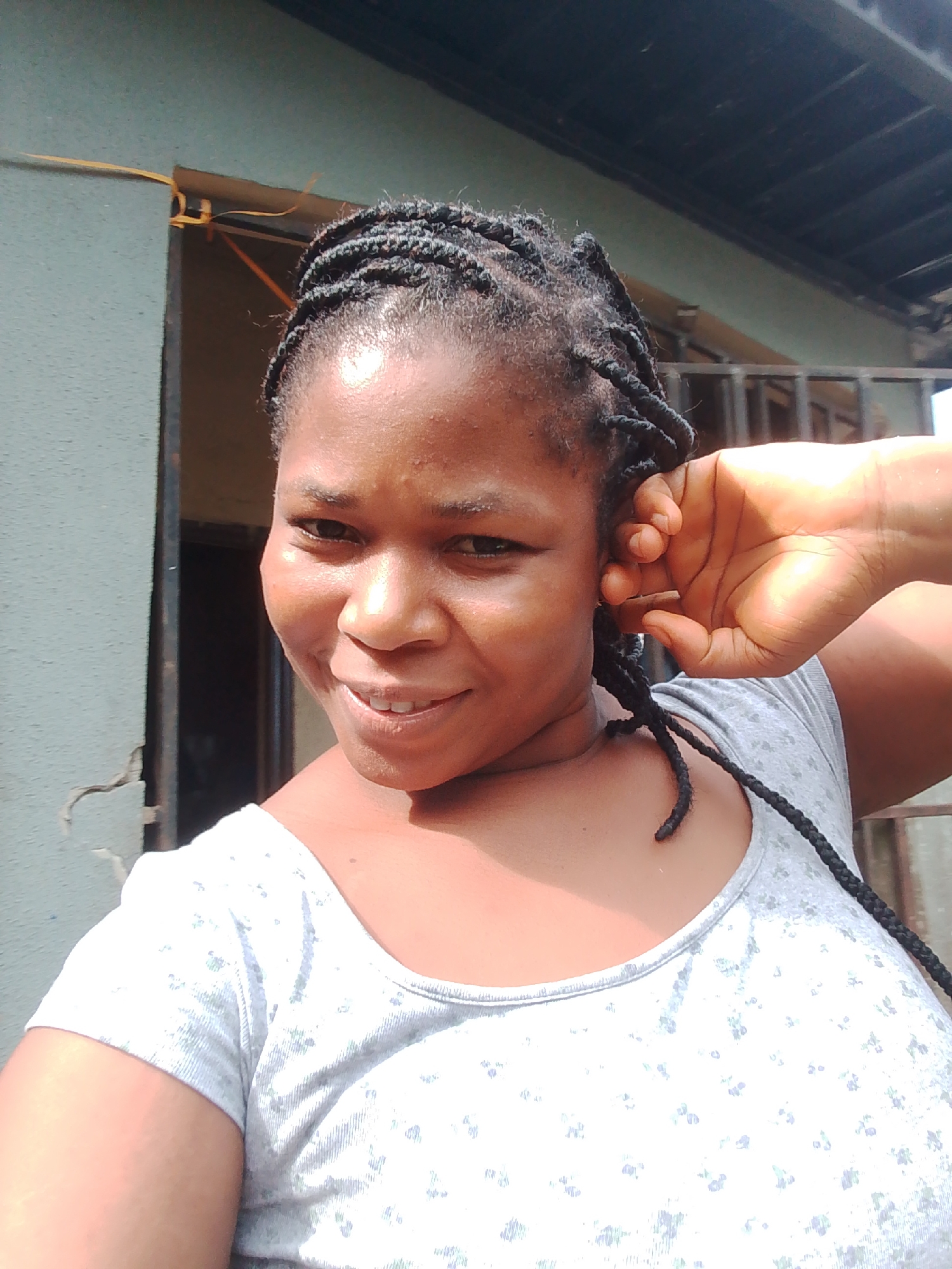A medium shot shows a young Black woman with braided hair from the shoulders up. She is looking directly at the camera with a slight smile. Her right hand is raised to her head, with her fingers touching her ear. She is wearing a light gray t-shirt with a subtle floral pattern. The background appears to be outdoors, with a weathered wall on the left and a corrugated metal roof structure above. The lighting suggests it is daytime, possibly sunny. The scene has a casual, everyday feel. There is no visible text in the image.