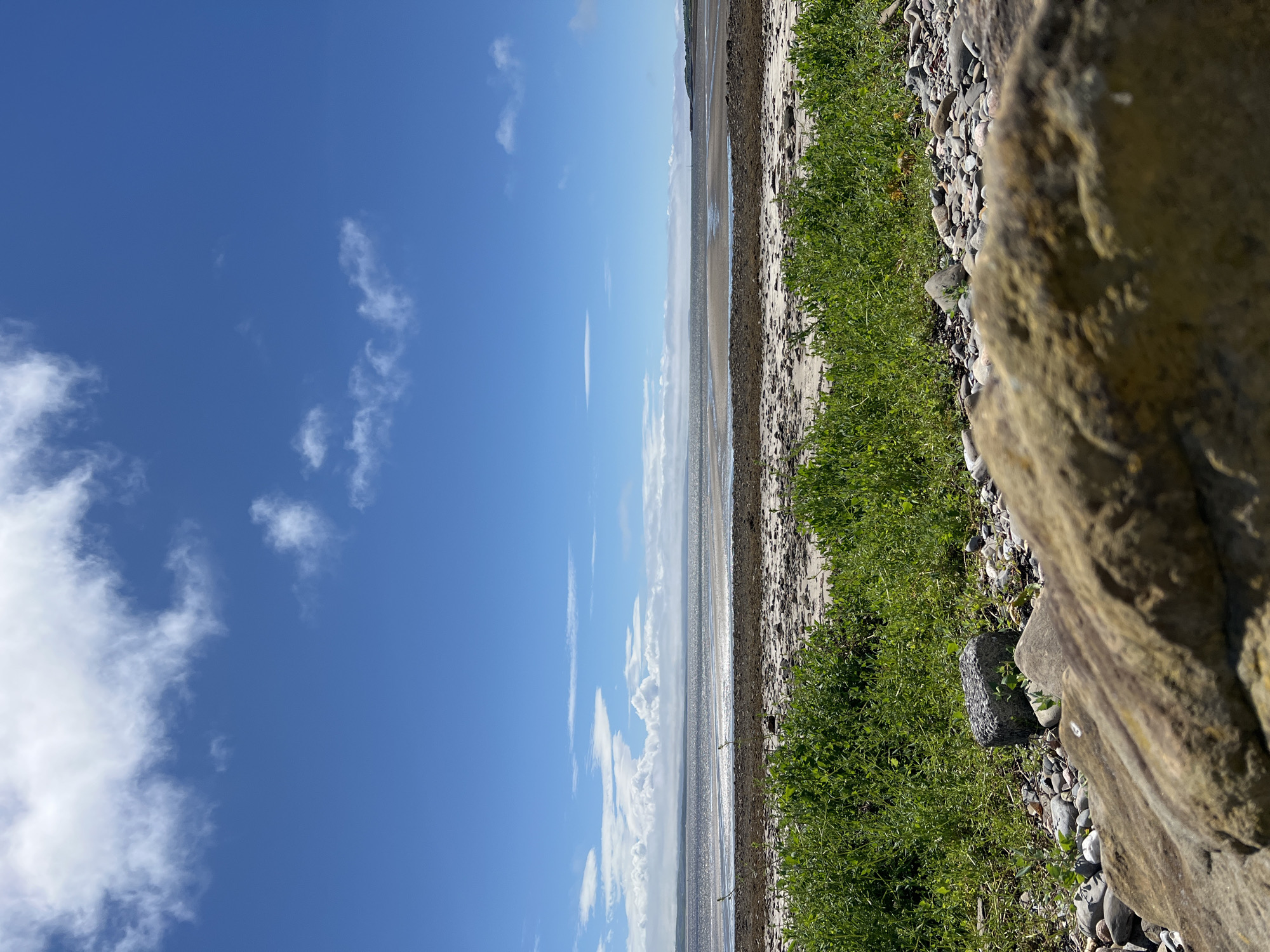 The image depicts a coastal scene with a bright blue sky filled with scattered white clouds. A body of water, likely the ocean, stretches out towards the horizon, appearing shimmering and distant. The foreground is a rocky shoreline covered in vibrant green grass and scattered stones. A large, textured rock is prominently featured in the lower right corner of the frame, adding a sense of depth and texture to the scene. The sun appears to be high in the sky, suggesting it is daytime, and the weather seems pleasant with clear skies and sunshine. There are no people or man-made objects visible in the image. The overall mood is serene and natural, capturing the beauty of the Irish coast.