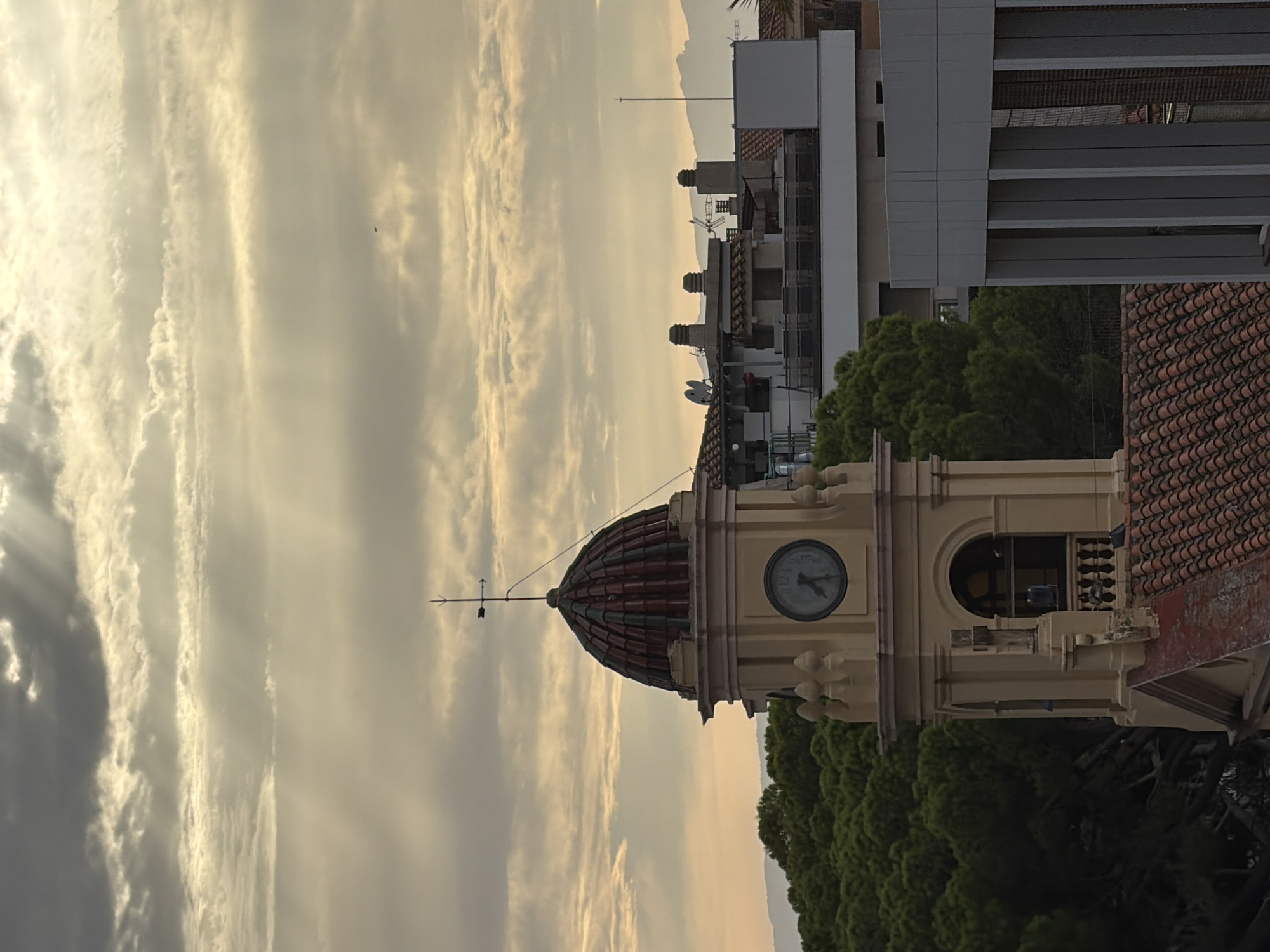 The image shows a view of the city of Salou, Spain, with a clock tower in the foreground. The sky is cloudy with sunbeams breaking through, suggesting either sunrise or sunset. The clock tower, with its tiled dome and clock face, is the central subject. In the background, there are other buildings, including some with tiled roofs and modern architectural elements. A dense cluster of green trees, likely pine trees, is visible to the left of the clock tower. The overall scene depicts an urban landscape with a touch of natural elements, under a dramatic sky.