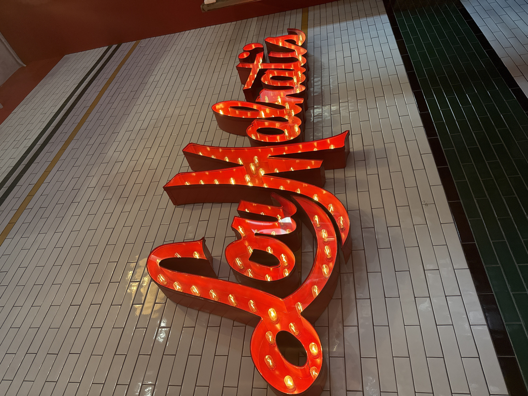 A sign reading "Smoque" in large red illuminated letters is mounted on a white-tiled wall. The wall features two vertical stripes, one dark green and the other yellow. The location is most likely a Smoque BBQ restaurant in Chicago.