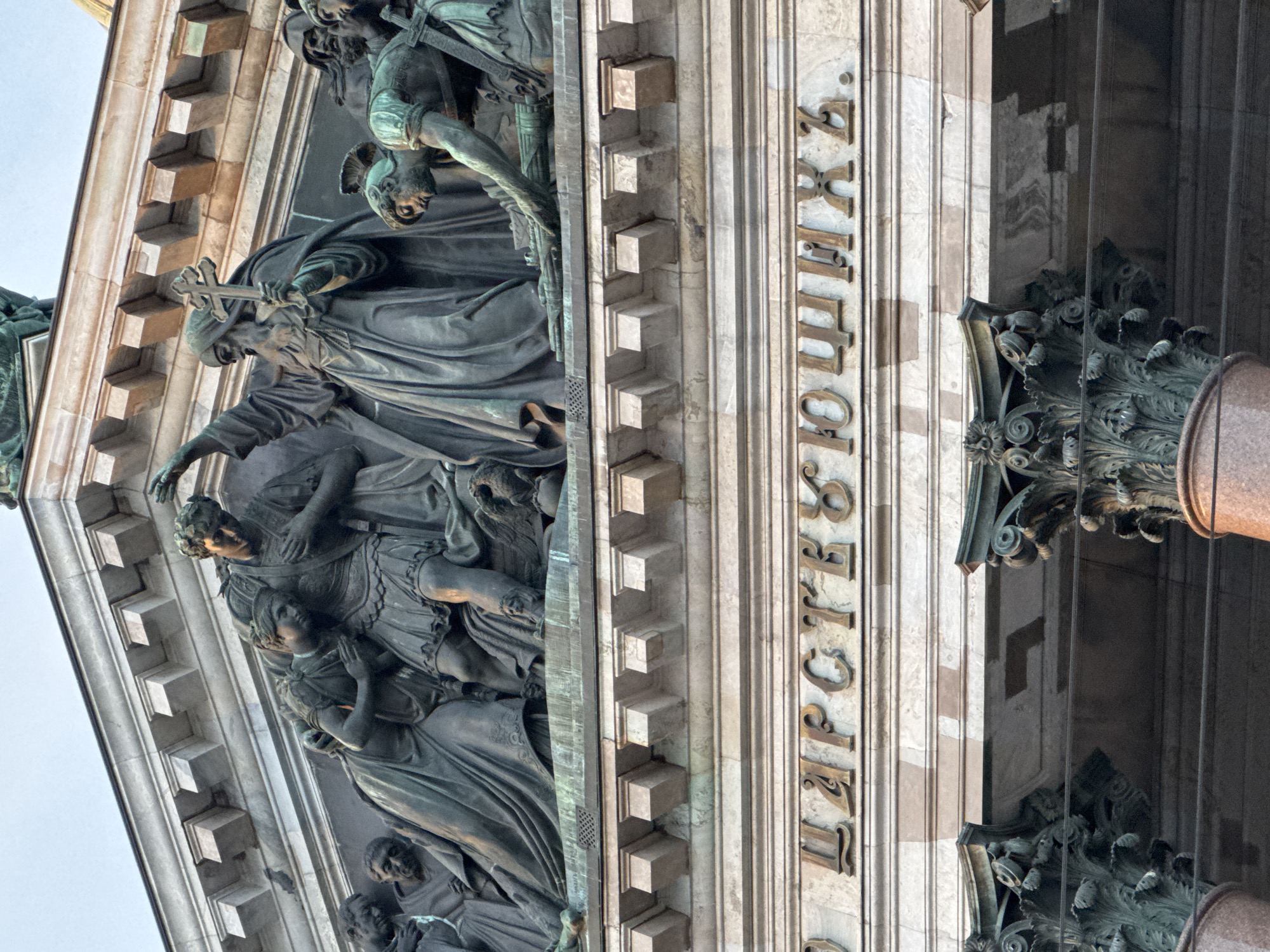 The image shows a close-up, low-angle view of a section of the facade of St. Isaac's Cathedral in Saint Petersburg, Russia. The upper portion of the image features a relief sculpture depicting several figures in bronze. A central figure, possibly a religious leader, is raising a cross with his right hand. To his left, two figures are embracing, one with a hand on their chest and the other with an arm around them. To the right of the central figure, another person is kneeling and appears to be handling an object. The figures are detailed and appear to be made of aged bronze, with some green patination visible.
Below the relief sculpture is a band of marble with an inscription in Cyrillic letters. The letters are golden and prominently displayed. The inscription reads "ЦАРСТВУЮЩИХЪ" (Tsarstvuyushchikh).
Further down, the image shows the upper parts of two ornate Corinthian columns. These columns are also made of stone, possibly marble, and have intricate carvings. The architectural style suggests a neoclassical or Beaux-Arts design. The background is a soft, pale sky. The overall impression is one of grandeur and historical significance.