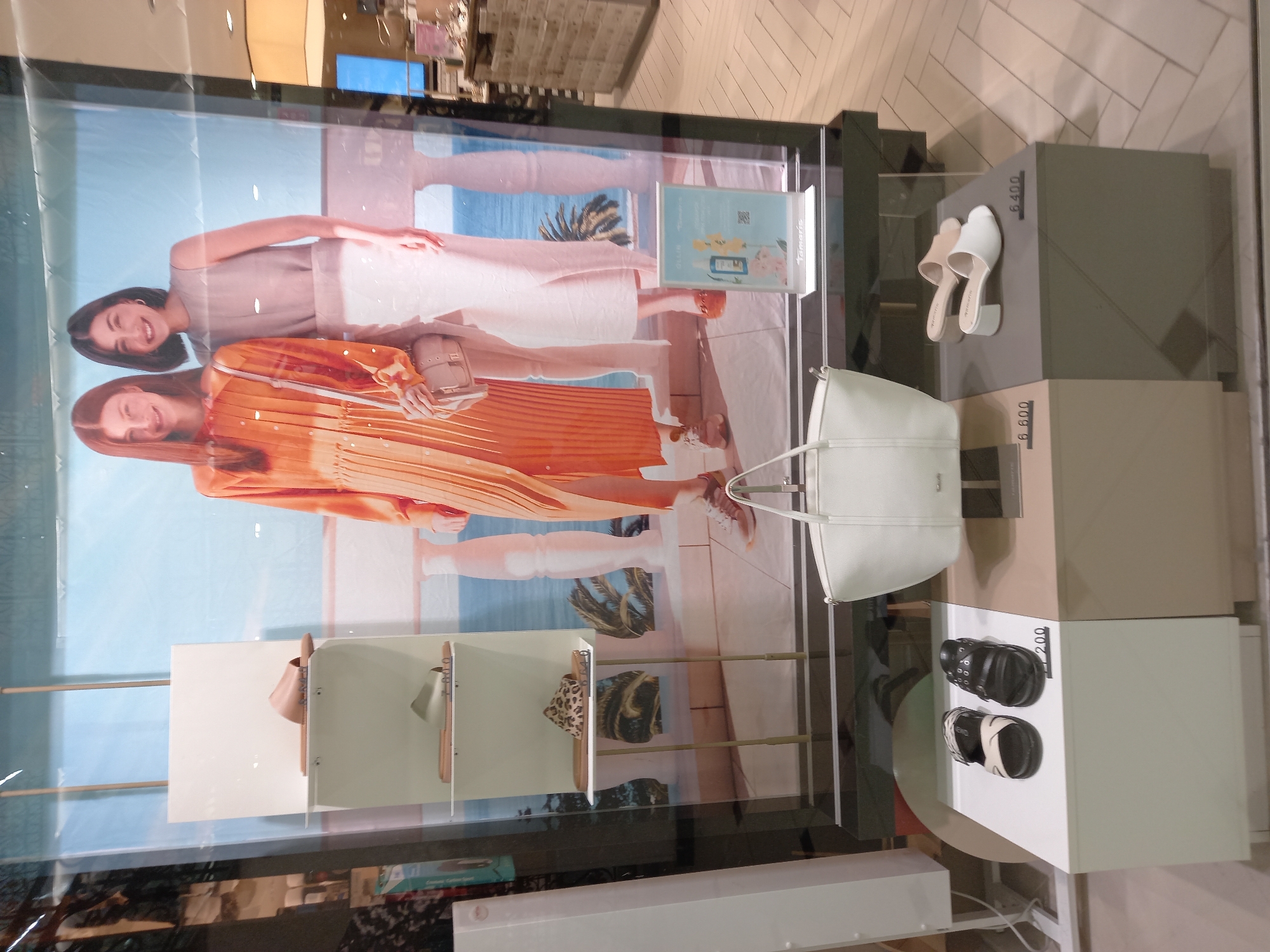 The image displays a retail store window featuring a display of women's shoes and a handbag. In the background, a large poster shows two women in fashionable attire, with a backdrop suggesting a coastal scene. On the left side of the window, a white shelving unit presents several pairs of women's sandals and flip-flops. In the foreground, a white handbag is placed on a display platform. To the right of the handbag, two pairs of women's slip-on shoes are displayed on another platform, each with a price tag indicating 6,400. Below the handbag and to the left, another display shows two pairs of women's sandals with a price tag of 7,200. The floor of the store appears to be tiled in a herringbone pattern. The overall scene depicts a product display within a commercial establishment.
