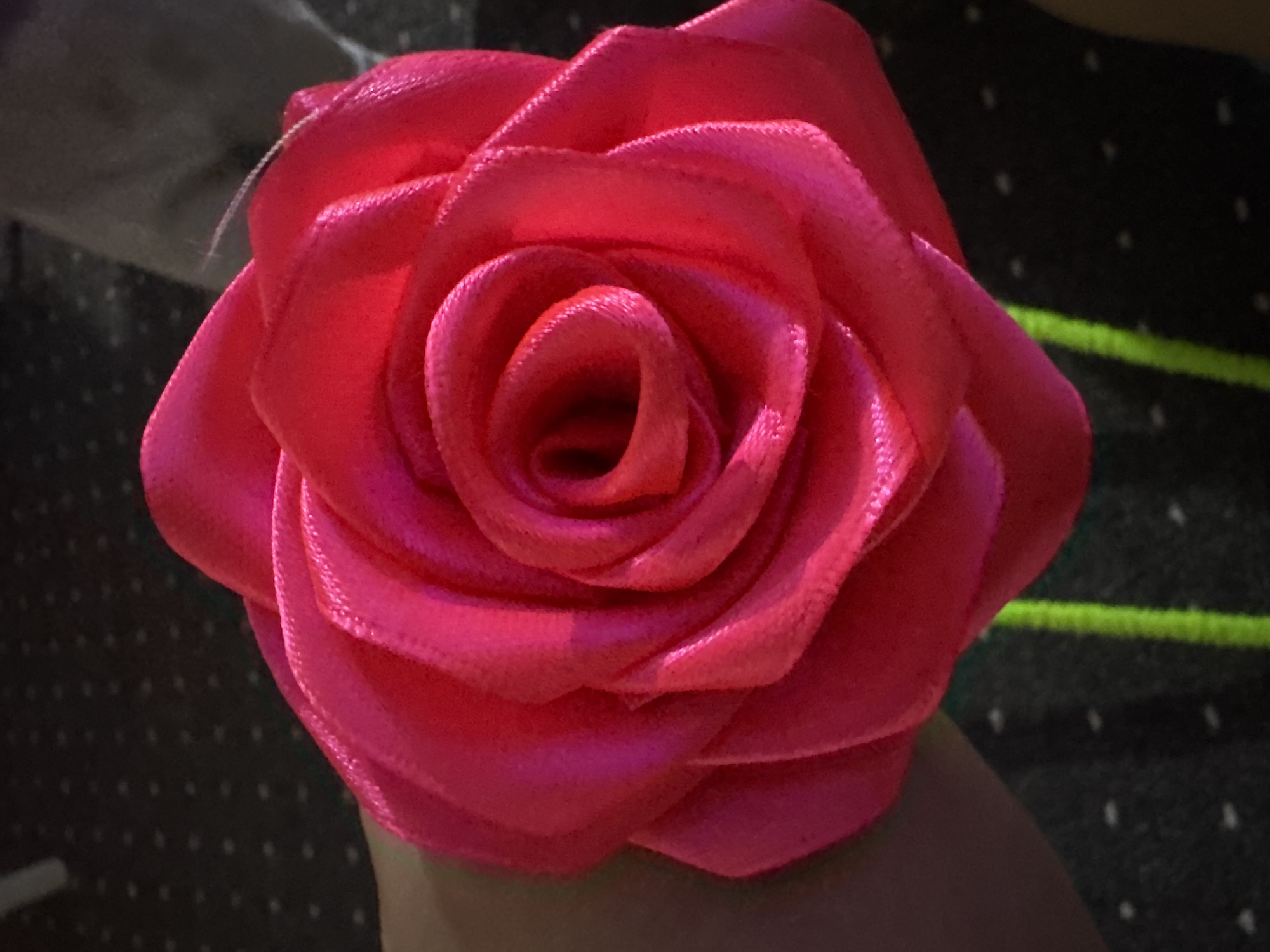 The image shows a close-up, overhead view of a bright pink satin rose. The rose is held in someone's hand, with a portion of their arm and hand visible on the left side of the frame. The rose is elaborately layered, with petals tightly curled towards the center, creating a spiral effect. The satin material has a slight sheen and texture. The background is dark and appears to be a patterned fabric, possibly a tablecloth or upholstery, with small, light-colored dots. Two thin, bright green stems or ribbons are visible below the rose, extending downwards and slightly out of focus. The lighting suggests an indoor setting. The location is not depicted and cannot be determined from the image alone, therefore no information about Chardara, Uzbekistan can be provided.
