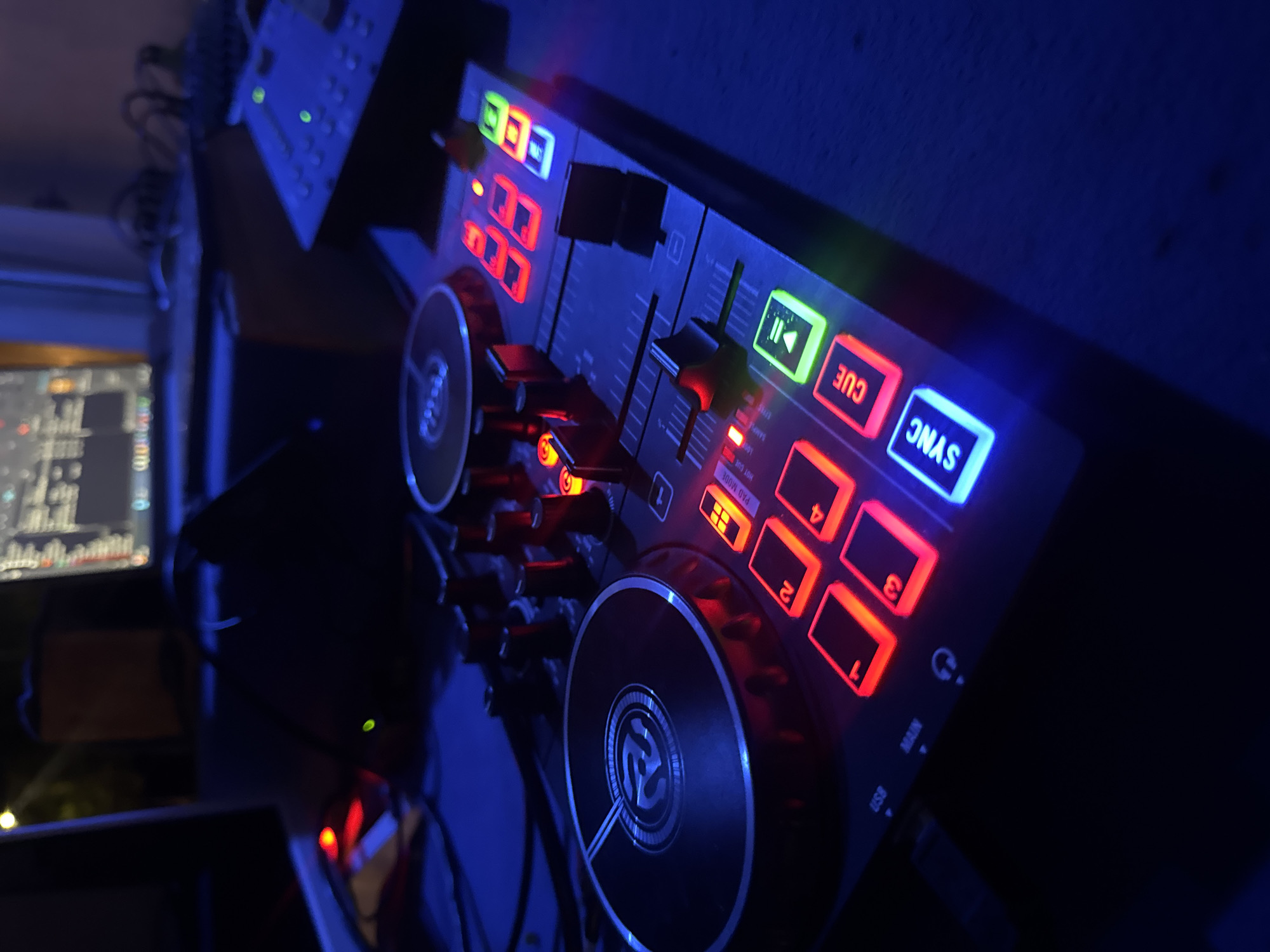 The image shows a close-up, low-angle, dimly lit shot of a DJ controller with illuminated buttons. The controller is black with a metallic jog wheel that has a logo. Several buttons are lit up in red, orange, and blue, including ones labeled "SYNC," "CUE," and numbers 1-4. In the background, a computer monitor displays a grid-like interface, suggesting a digital audio workstation. The overall scene is dark and atmospheric, with blue ambient lighting dominating the scene. The setting appears to be an indoor space, likely a home studio or a dimly lit entertainment area. The image does not contain any visual information that can be used to determine its location in the city of Isola delle Femmine, Italy.