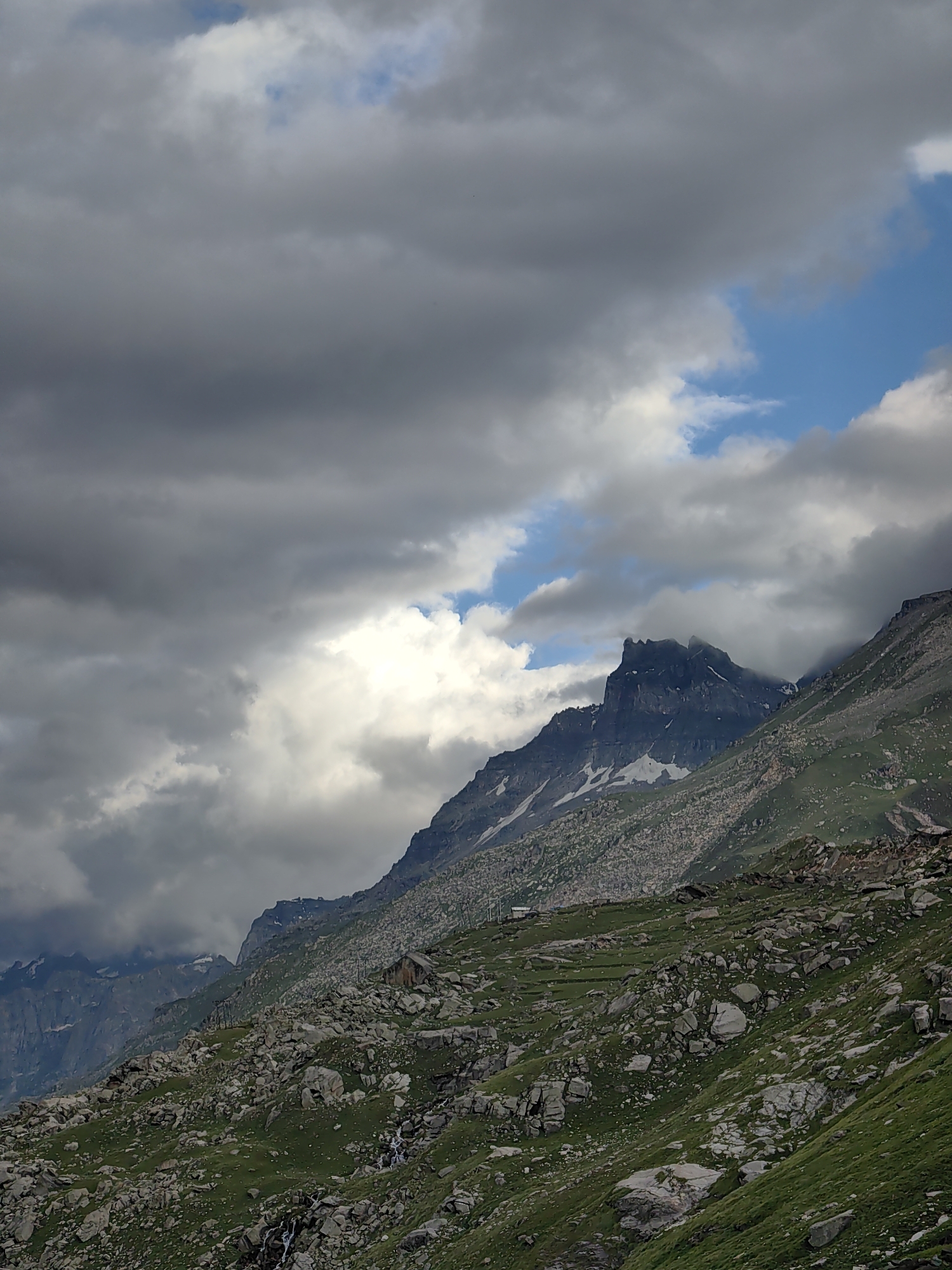 This image depicts a dramatic mountain landscape under a cloudy sky. The foreground is dominated by a rocky, grassy slope, with scattered boulders and patches of green vegetation. A small stream or waterfall is visible cascading down the rocks. In the mid-ground, a steep mountain range rises, characterized by rugged terrain, rocky outcrops, and patches of snow. One prominent peak in the center has a sharp, jagged summit. The upper portion of the image is filled with a heavy, overcast sky, with dark, gray clouds obscuring much of the blue sky, though a few patches of clear blue are visible, along with bright white clouds illuminated by the sun. The overall impression is one of a wild, untamed, and somewhat moody natural environment. The location is identified as Yari-Khoksar, India. There are no people or animals visible in the image.