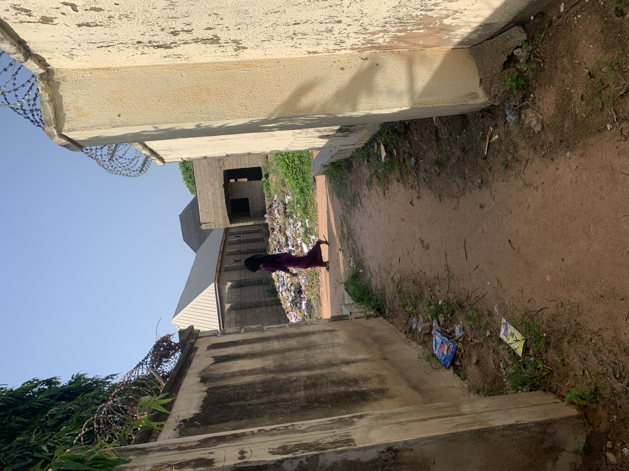 In Jalingo, Nigeria, a young woman with long dark hair, wearing a long purple dress, walks along a dirt path between two walls. The wall on the left is concrete and stained, while the wall on the right is stuccoed and weathered. Barbed wire fences top both walls. Beyond the walls, a large pile of trash is visible, with construction debris and what appears to be more unfinished buildings in the background. The ground is dry and dusty, with sparse patches of green grass and weeds growing along the edges of the path and walls. Several pieces of litter, including wrappers, are scattered on the ground. The sky is clear and blue.