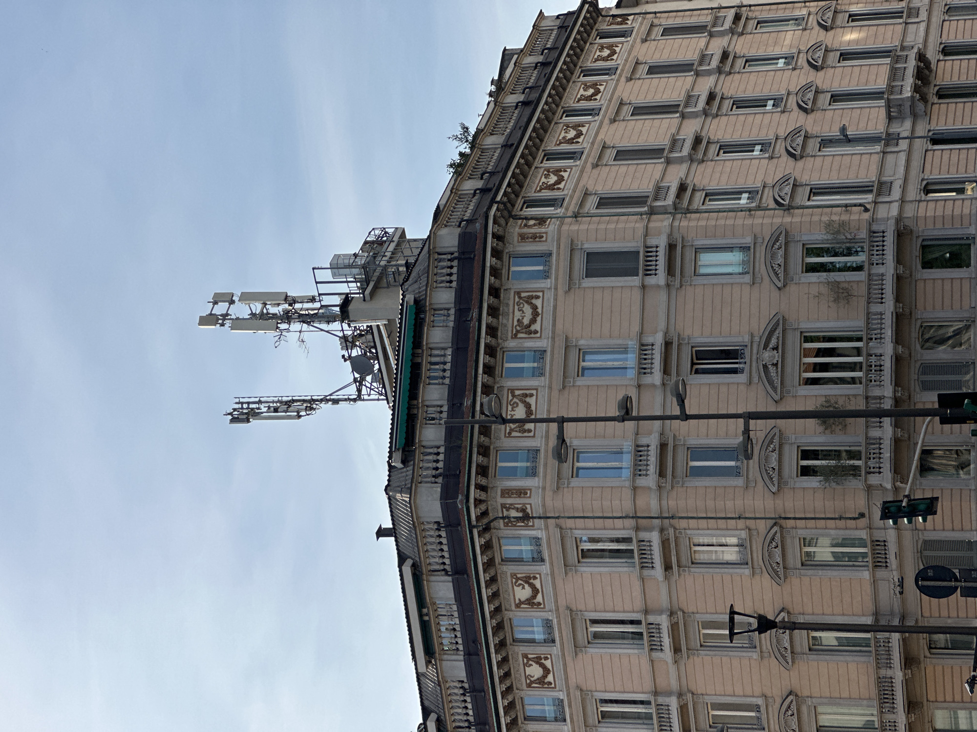 This is an exterior, slightly low-angle photograph depicting the upper floors and rooftop of a multi-story classical-style building in Milan, Italy. The building features a light salmon-colored stucco facade accented with light gray stone trim and ornate architectural details.
The upper main floor of the building is characterized by a series of rectangular windows, each surmounted by elaborate decorative panels depicting festoons and scrolls in a darker, contrasting color. Below these, a prominent cornice separates this level from a setback rooftop structure. This rooftop level includes a dark, possibly slate or metal, mansard-style roof section, topped by a stone balustrade that runs along the visible edges. Small patches of green foliage, possibly small trees or shrubs, are visible growing behind this balustrade on the right side.
Dominating the rooftop are two large, complex cellular antenna arrays or masts. These structures consist of multiple rectangular panel antennas, dishes, and associated cabling, all mounted on metal frameworks. A smaller, enclosed structure, possibly an equipment shelter or HVAC unit, is situated between the two main antenna arrays. A single, small black chimney is also visible on the darker roof section.
Further down the building, multiple floors are visible, each with a consistent pattern of rectangular windows. Many windows are equipped with small balconies featuring gray stone balustrades. Some windows have external shutters, with a few visible in a closed position. Reflecting the sky, some windowpanes appear light blue. On the lower visible balconies, small potted plants or trees are discernible.
In the foreground, two black street lamp posts extend vertically, partially obscuring the building's facade. The left lamp post has multiple fixtures. At the bottom left, a dark green traffic light signal and a partial blue circular road sign are visible. The sky above is light blue with some faint, wispy clouds. No people or vehicles are visible in the frame.